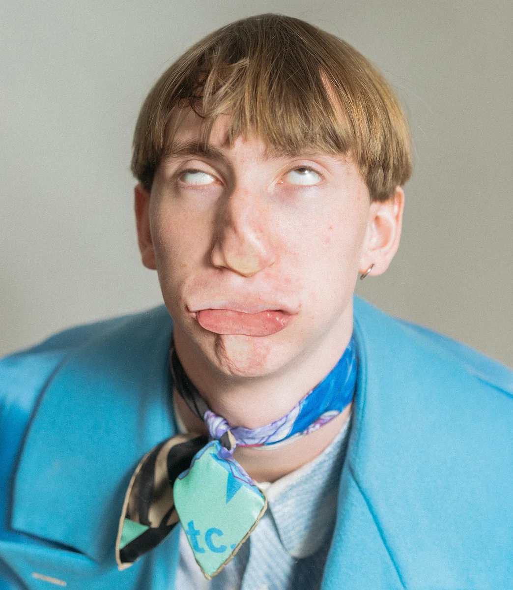 Model in a cyan coat and silk scarf from PSEUDONYM Autumn Winter 2019, face compressed against a rigid barrier for the Under Pressure campaign.
