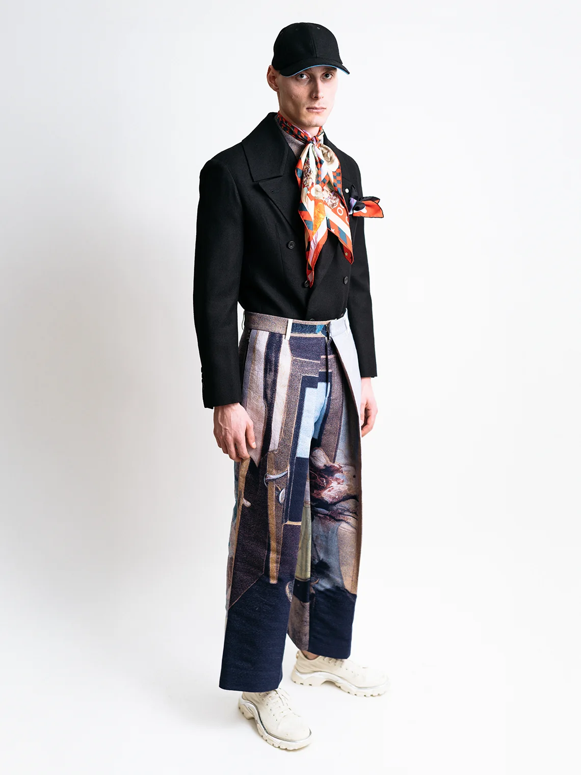 Model wearing a tucked black tailored jacket, silk scarf, black cap, and pleated jacquard trousers for PSEUDONYM Autumn Winter 2019.