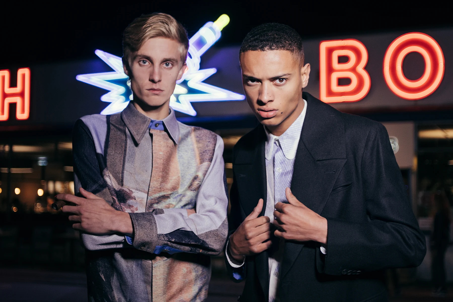 Two models wearing PSEUDONYM Autumn Winter 2019 against neon signage, featuring a printed silk panelled shirt and black tailored outerwear.