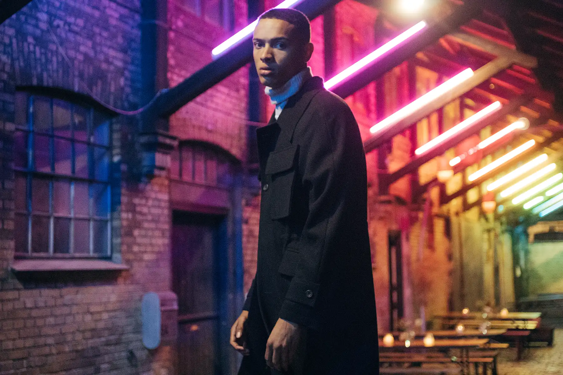 Model wearing black field coat over a cropped silk knit top under neon lighting for PSEUDONYM Autumn Winter 2019.