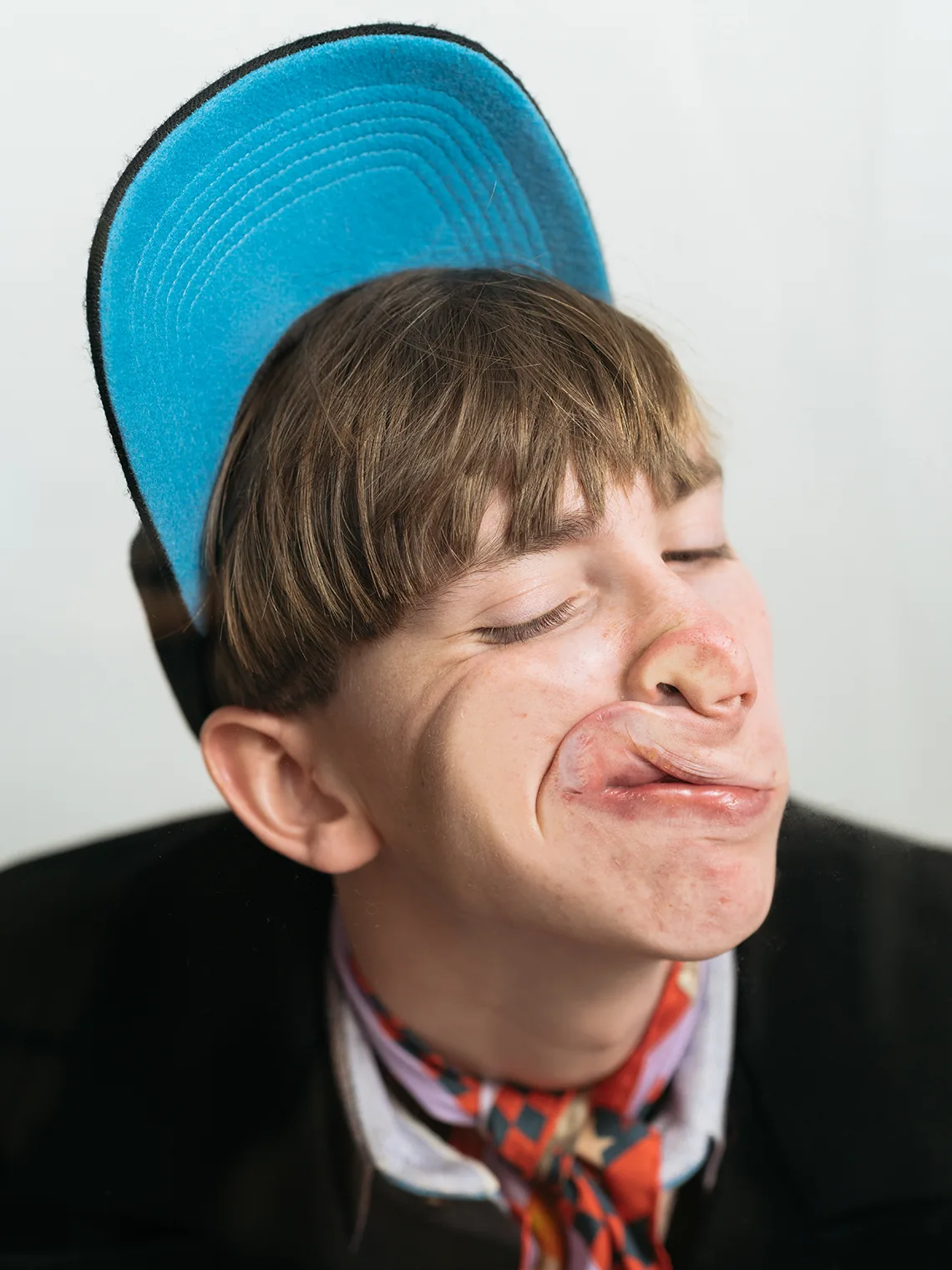 Model in a blue cap, silk scarf, and black double-breasted jacket, face compressed against a rigid barrier for the PSEUDONYM Autumn Winter 2019 Under Pressure campaign.