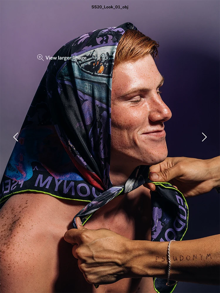 A smiling male model has a printed scarf tied around his head by another pair of hands.