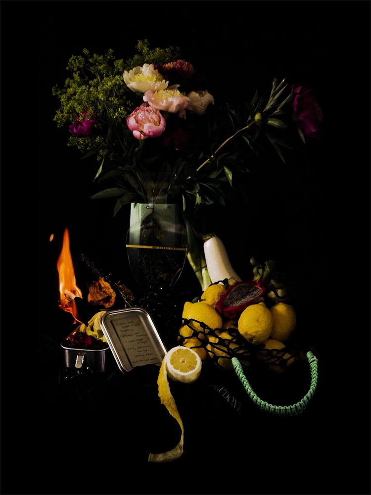 Dark still-life scene with flowers, fruit, vegetables, a small flame, and a black mesh bag with green handles.