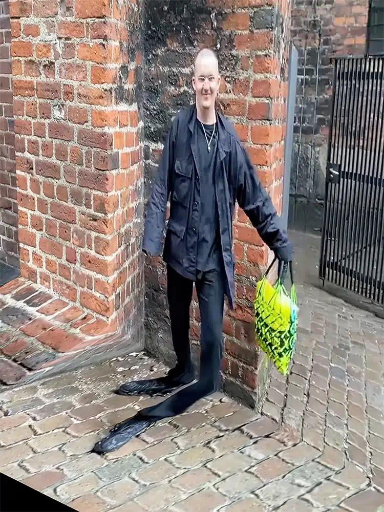 Man standing beside brick walls with warped perspective distorting his legs and the pavement.