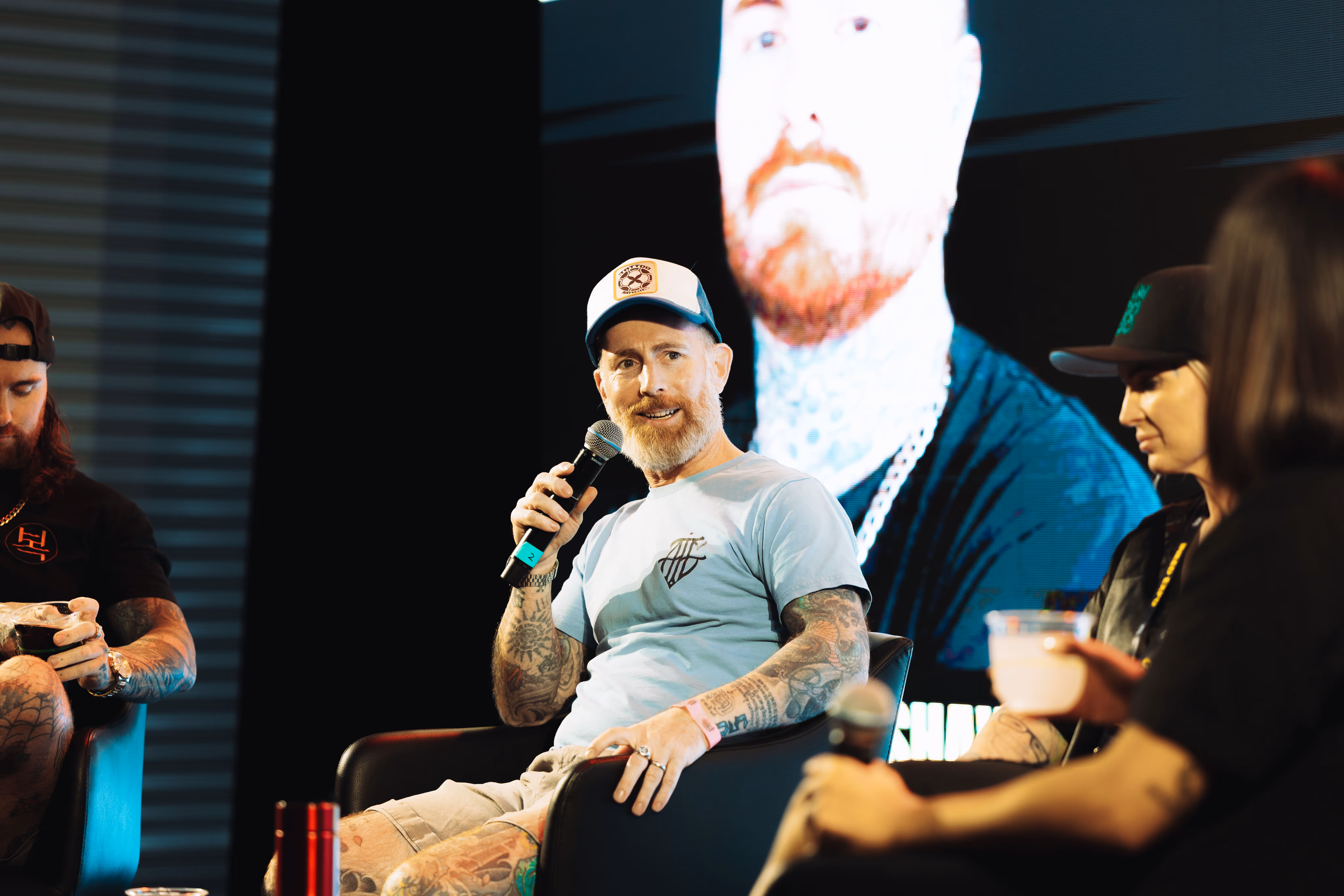 Man with tattooed arms wearing a light blue t-shirt and cap, speaking into a microphone while seated on a stage during a panel discussion.