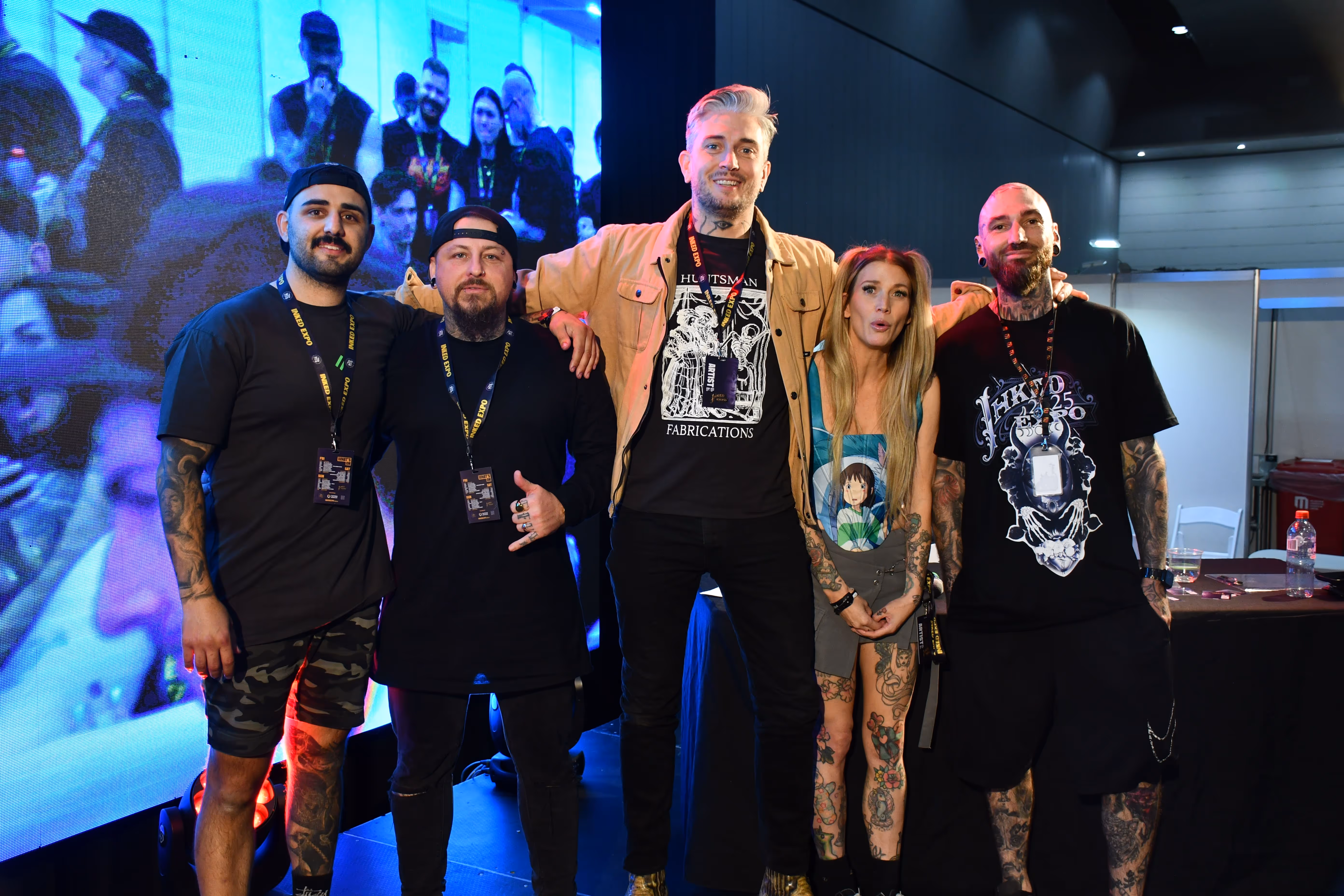Five tattooed people posing together at an indoor event with a large screen in the background.