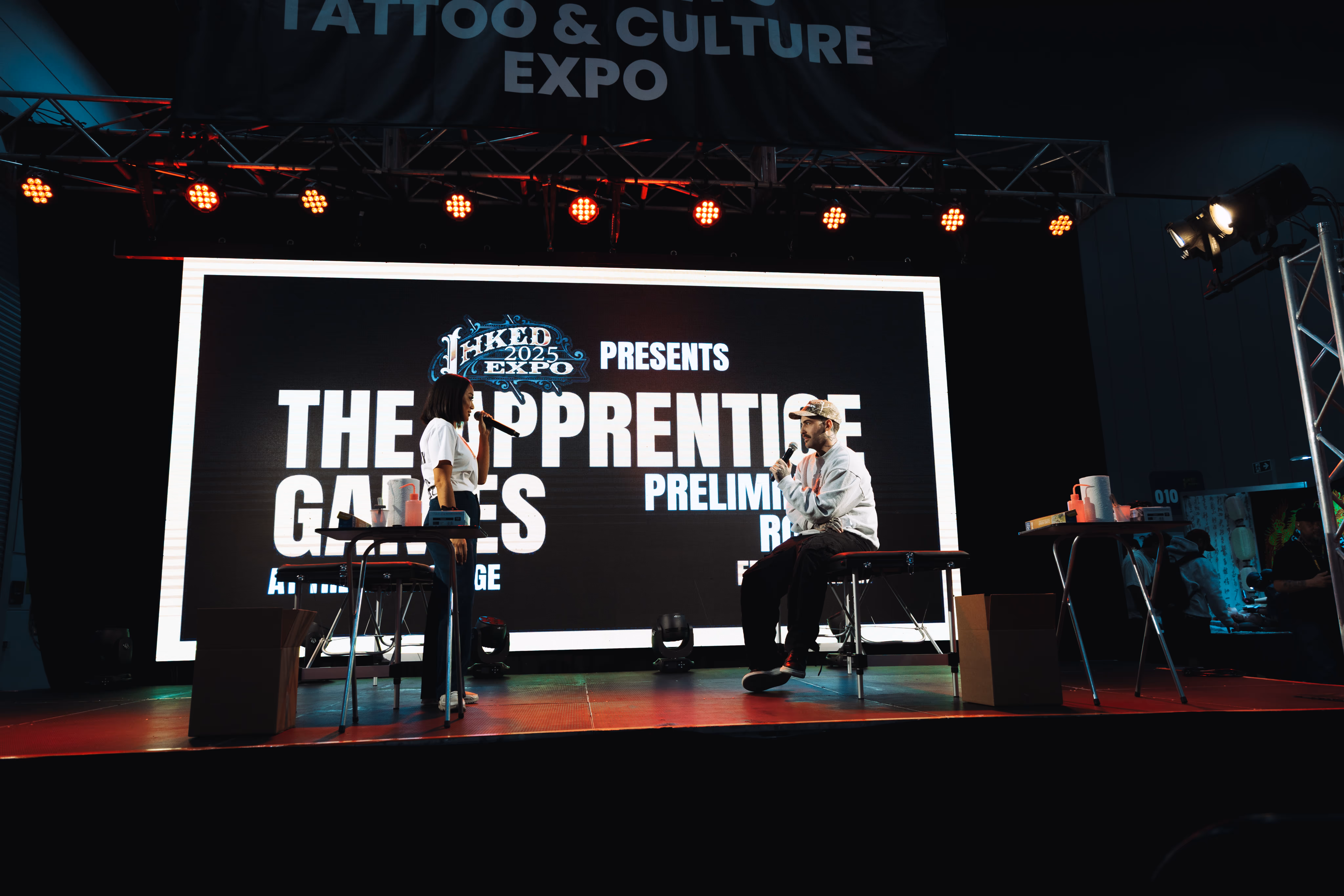 Two people on stage holding microphones with a large screen behind them displaying 'Inked 2025 Expo presents The Apprentice Games Preliminary Round'.