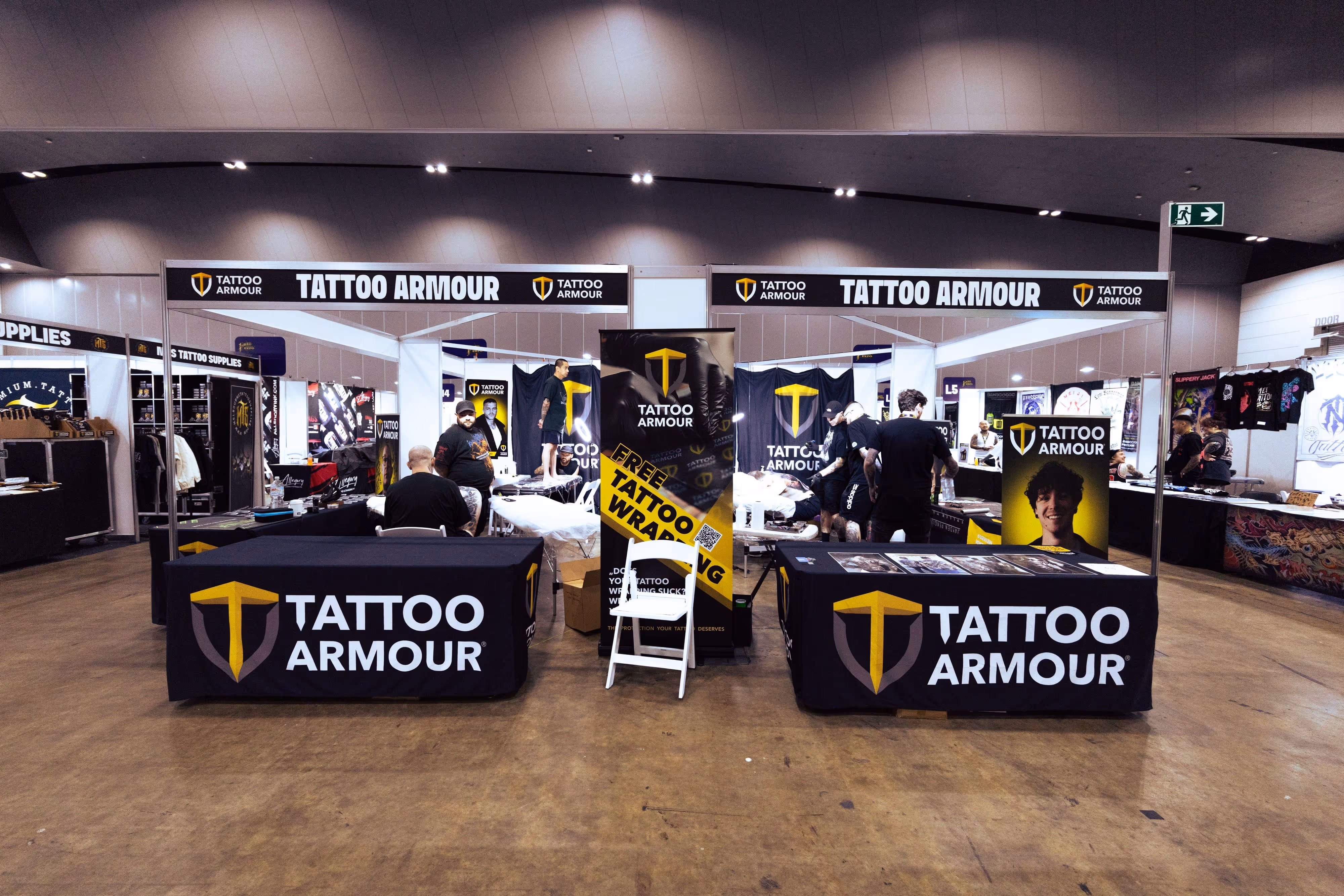 Tattoo Armour booth at a convention with tables, banners, and several people engaged in tattooing and discussions.
