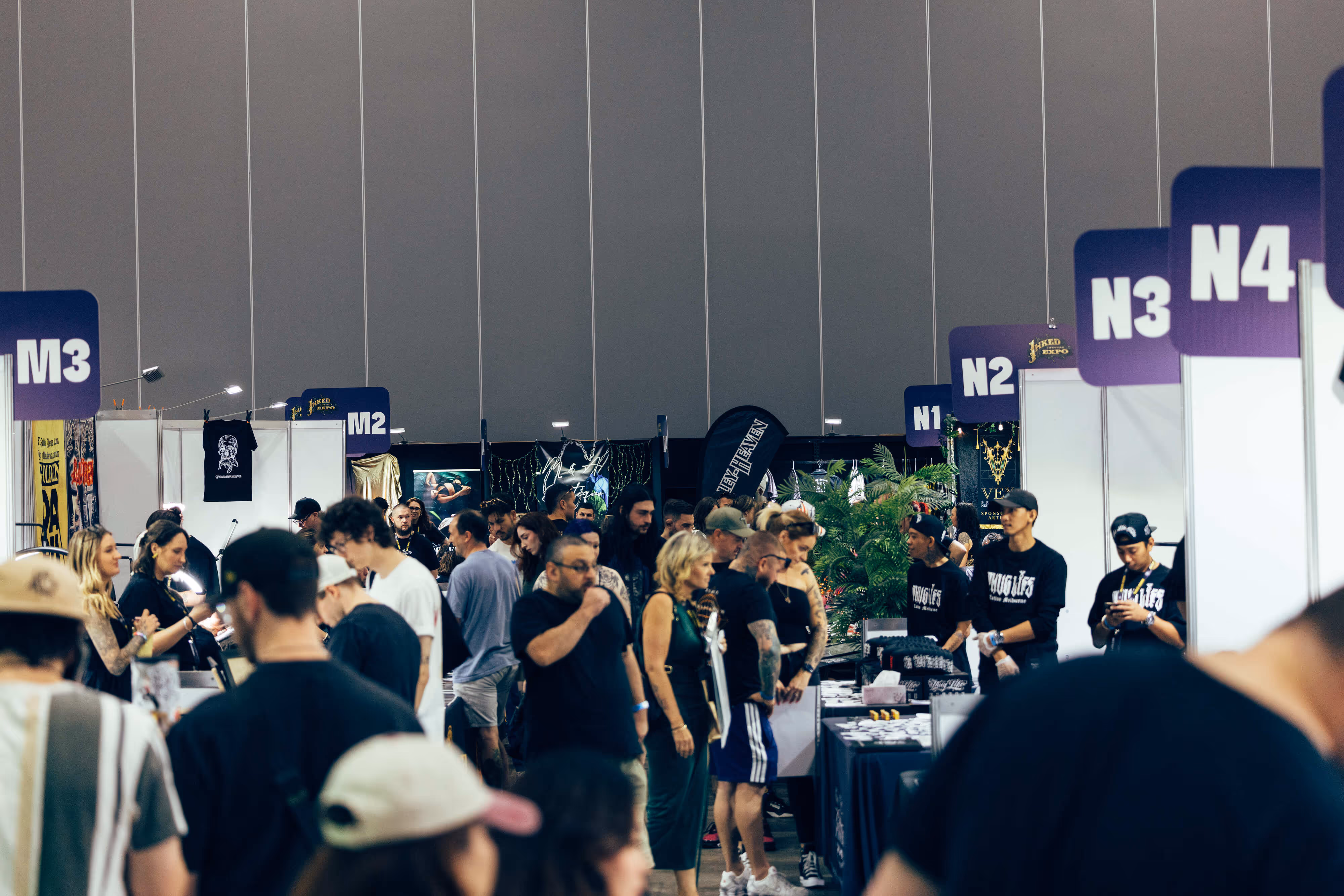 Crowd of people walking and browsing booths at a large indoor expo with numbered purple signs above each booth.