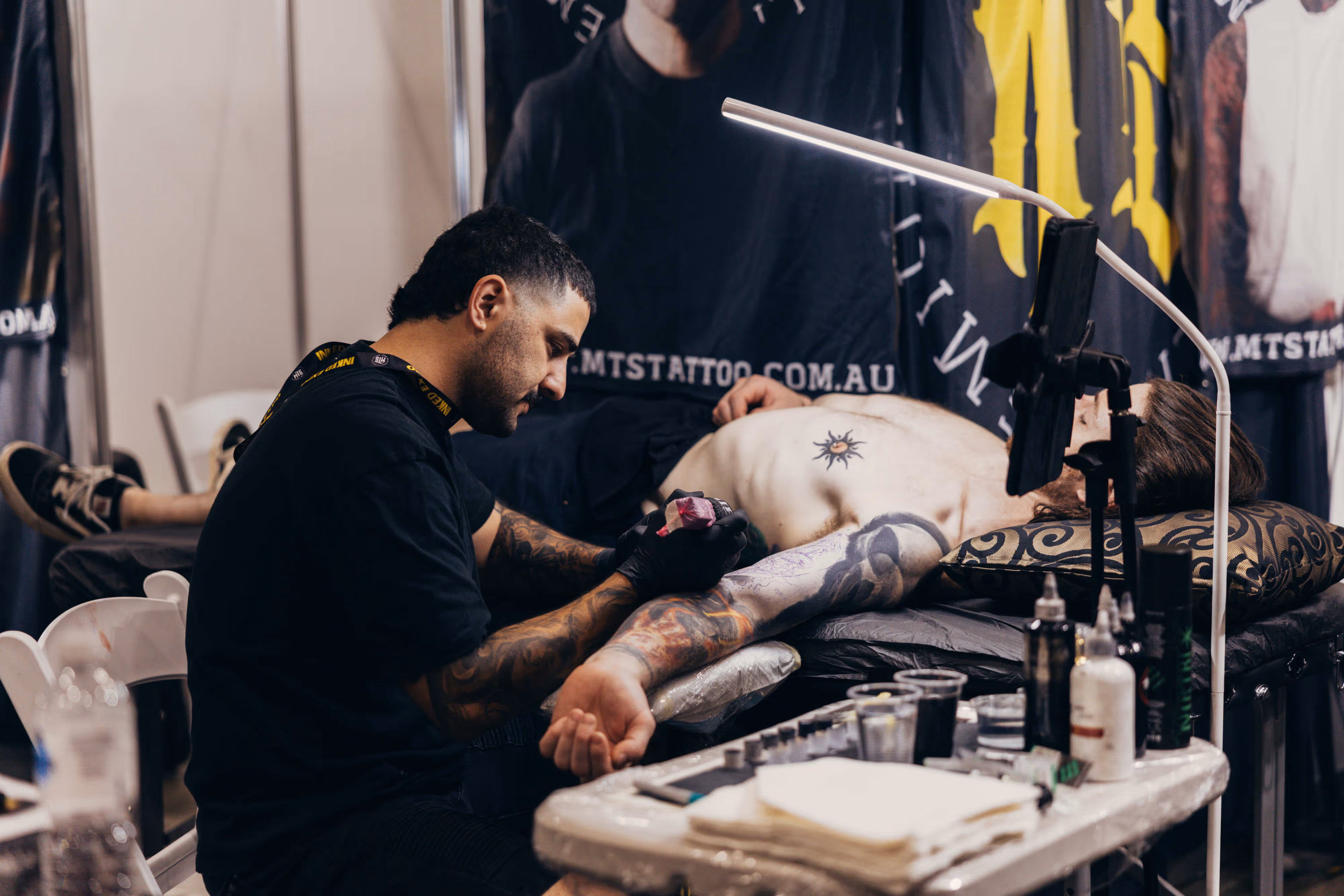 Tattoo artist in black gloves working on a man's arm tattoo while the man lies shirtless on a chair.