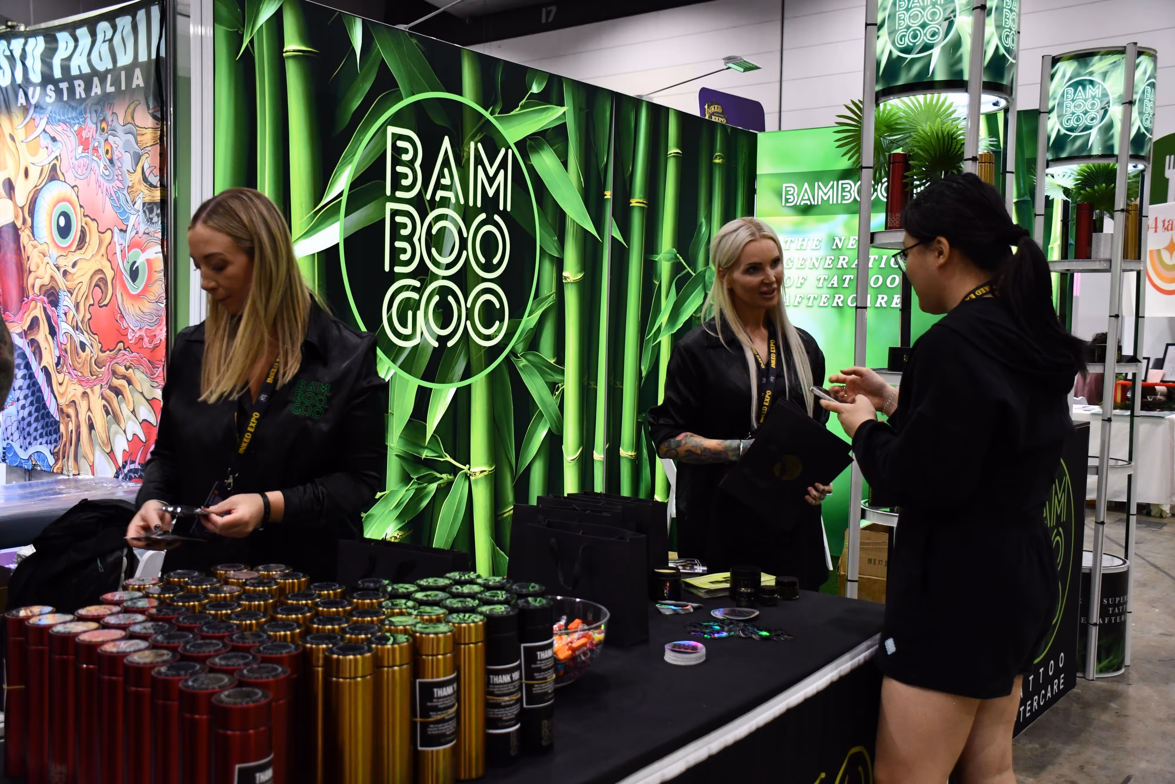 Three women at a Bamboo Goo booth with green bamboo-themed displays and products on a table at an expo.