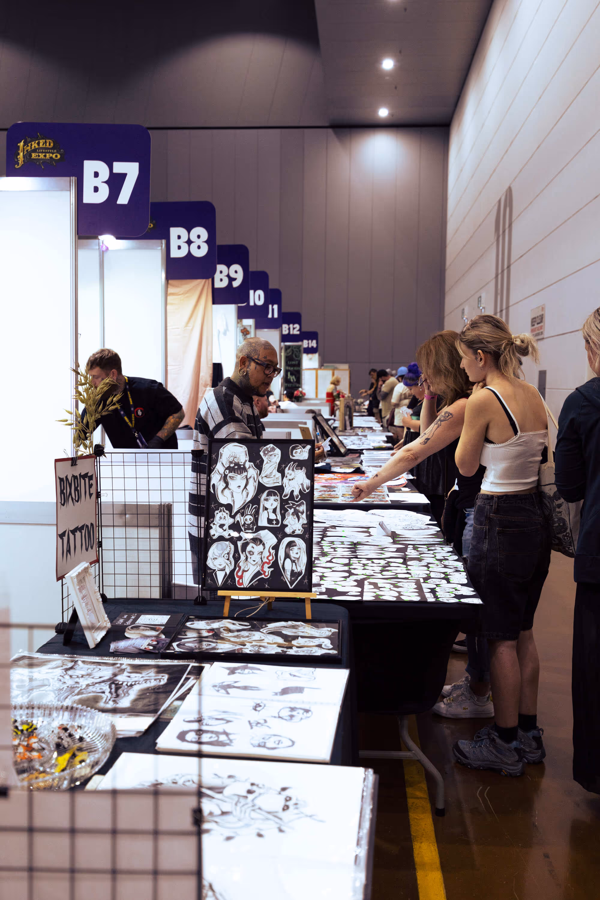 People browsing tattoo designs at booths numbered B7 to B14 in an indoor expo hall.