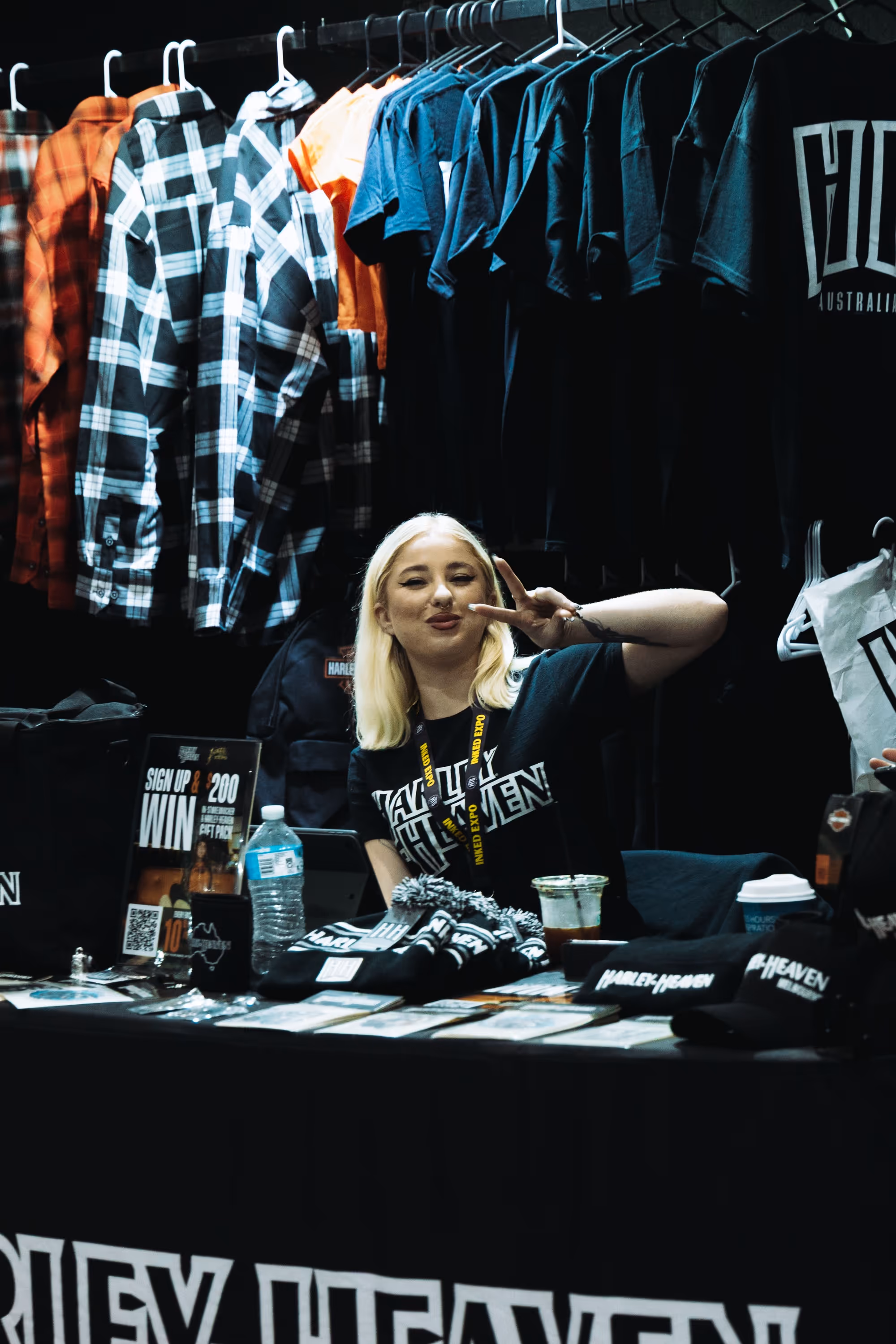 A smiling woman with blonde hair wearing a black Harley Heaven shirt poses with a peace sign at a merchandise booth with hats, shirts, and promotional materials displayed.