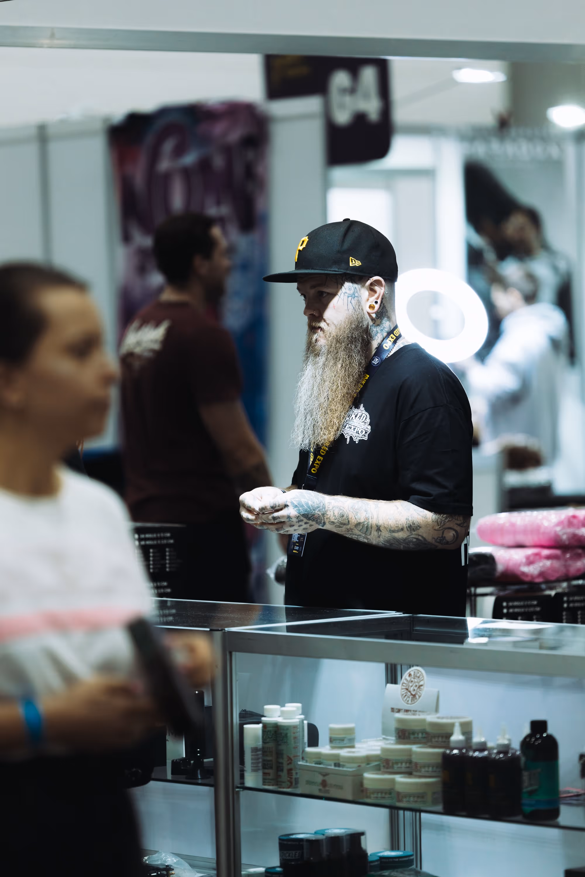 Tattooed man with a long beard and black cap standing behind a glass counter displaying skincare products, with other people blurred in the background.