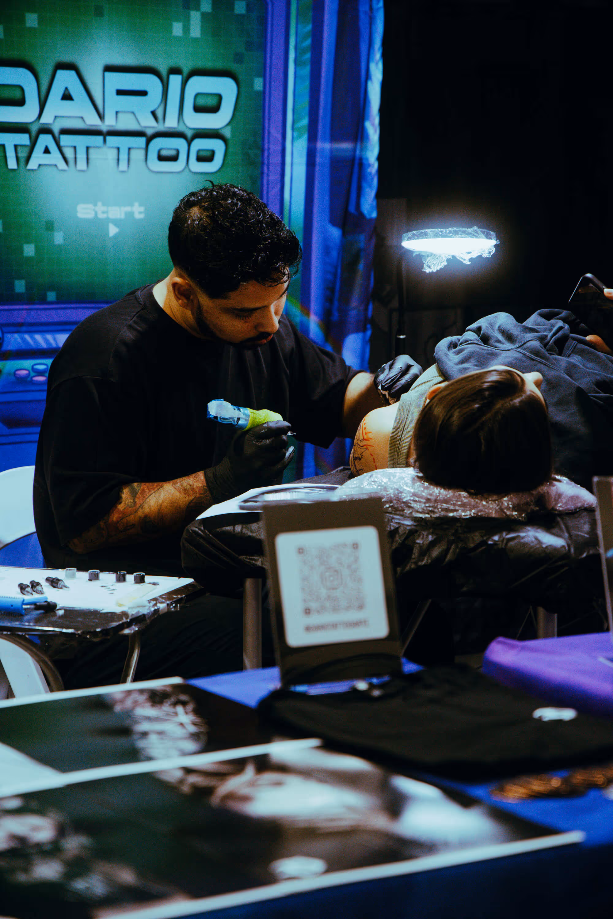 Tattoo artist wearing black gloves working on a tattoo on a client's shoulder in a studio with a green sign reading ‘DARIO TATTOO’ in the background.