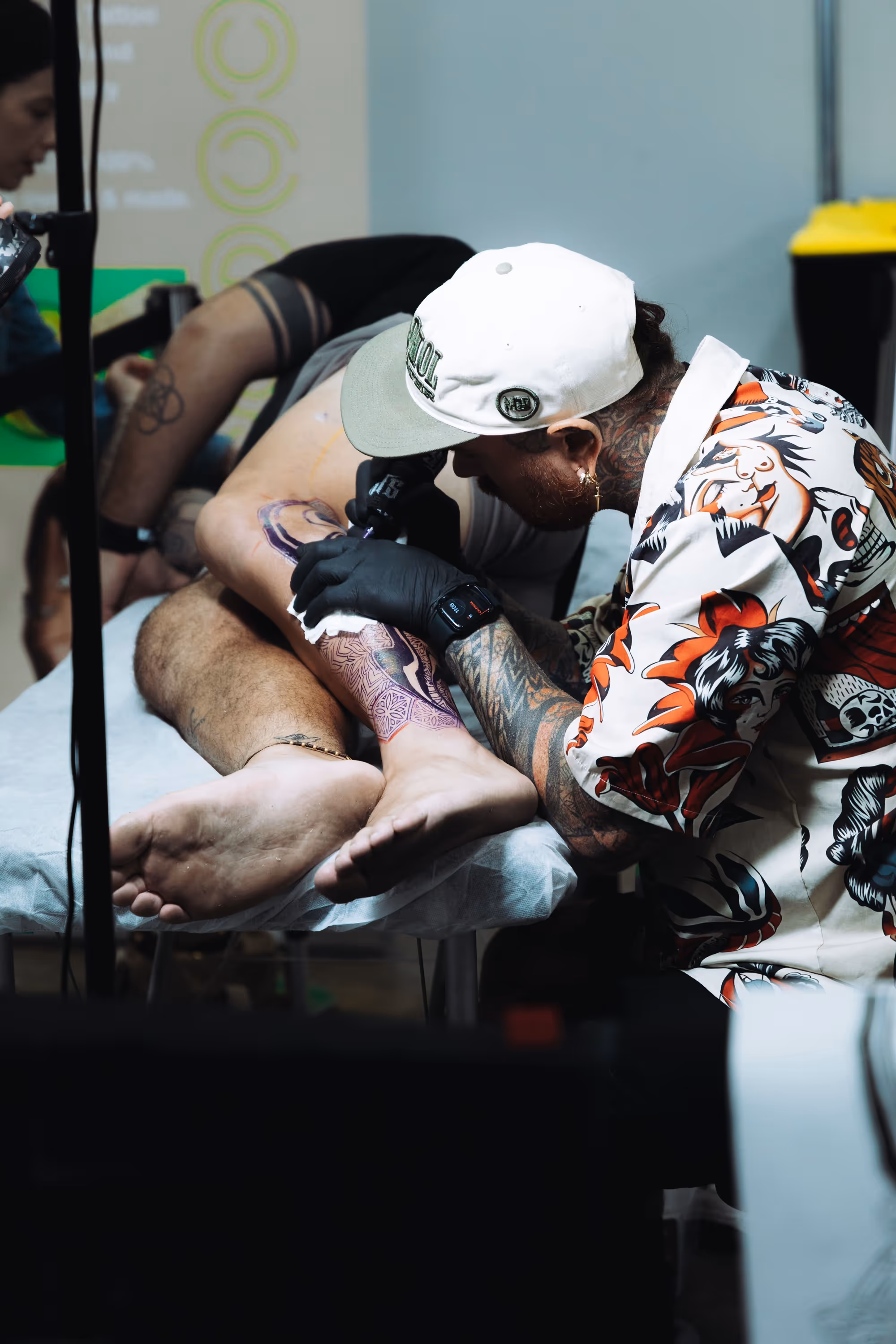 Tattoo artist with tattoos on his arms and wearing a patterned shirt inks a colorful leg tattoo on a client lying on a black and white table.