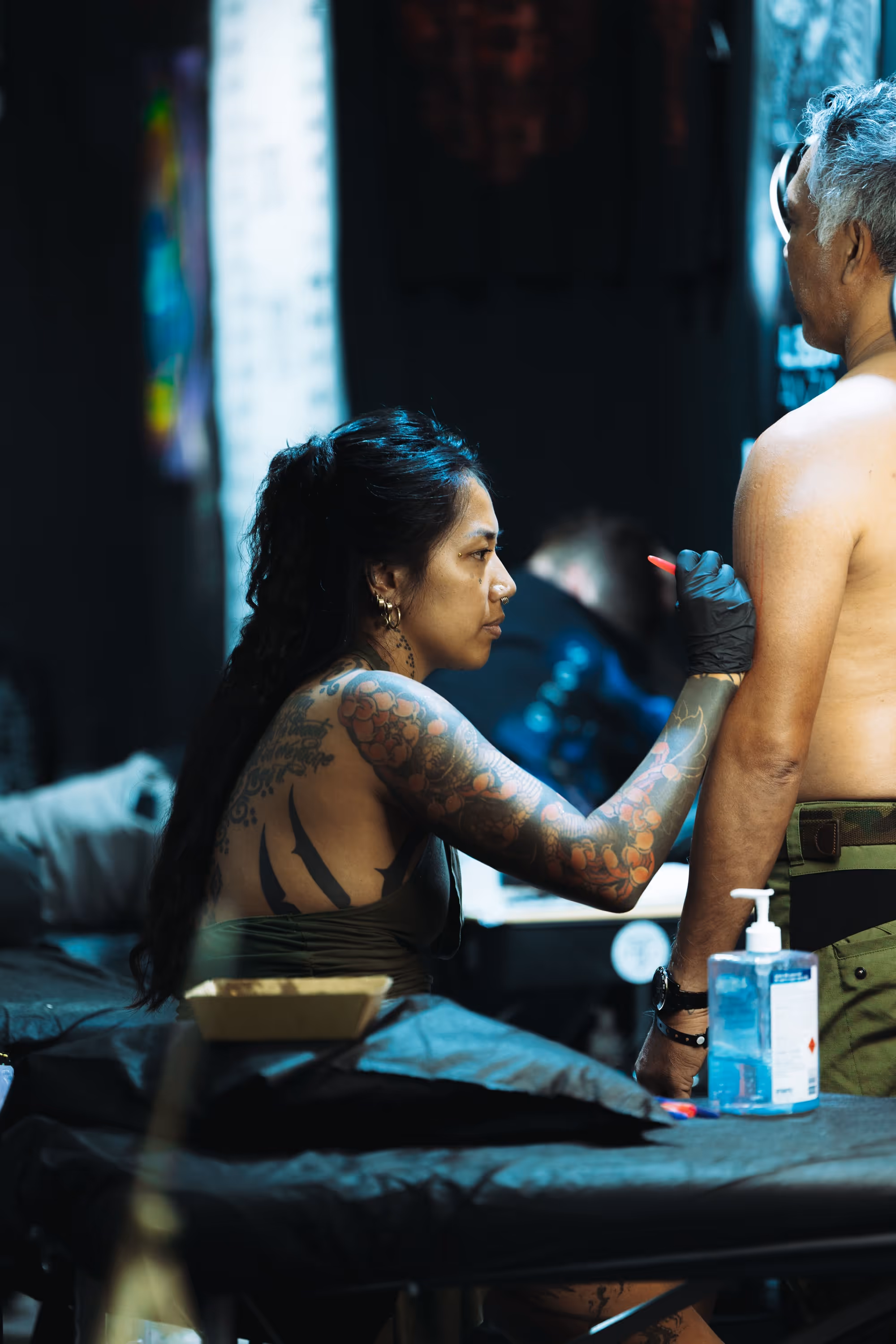 Tattoo artist with long black hair and sleeve tattoos preparing to ink a standing shirtless client in a dimly lit studio.