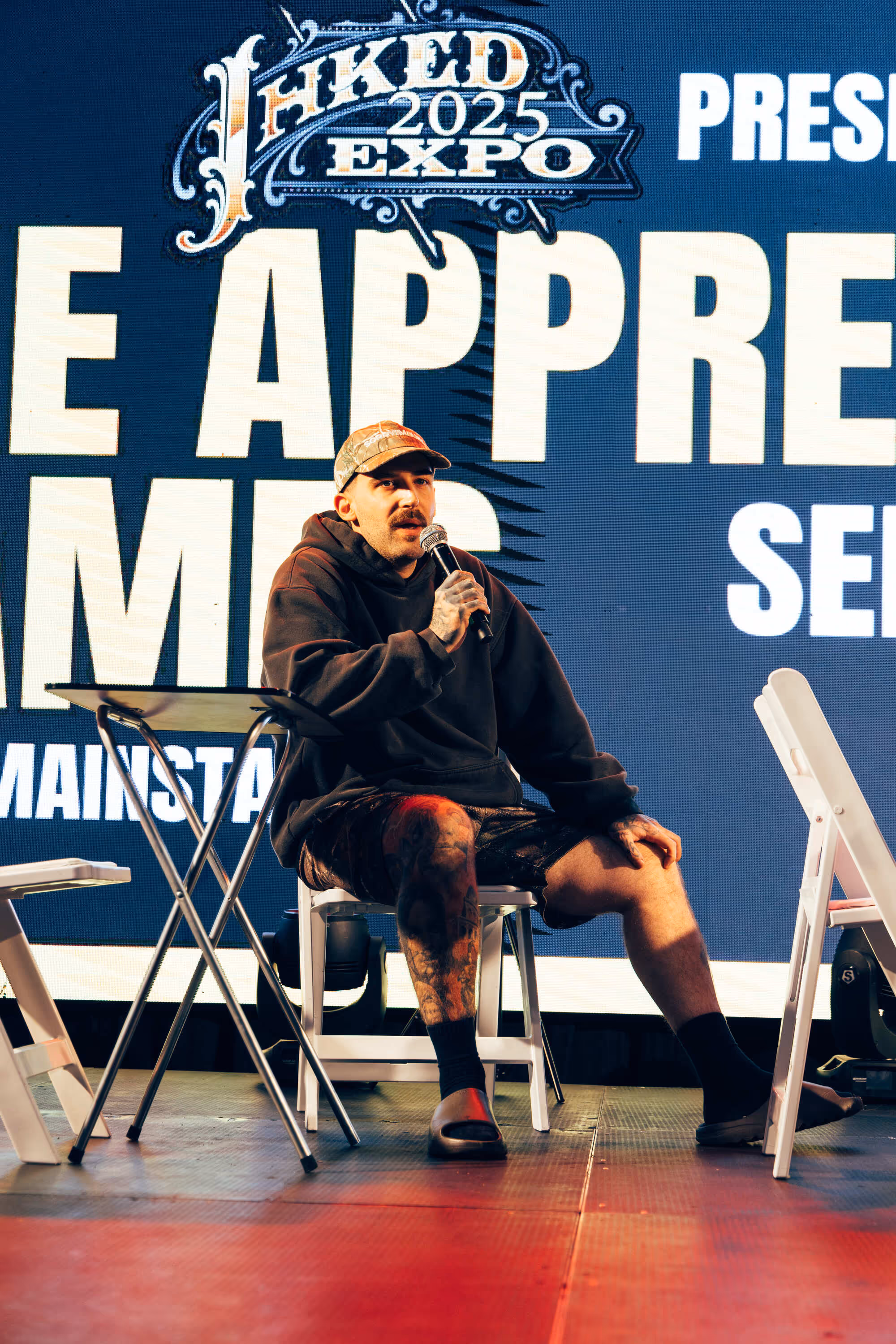 Man in a black hoodie, camo hat, and shorts with tattoos speaking into a microphone while seated on a white chair on stage at Inked Expo 2025.