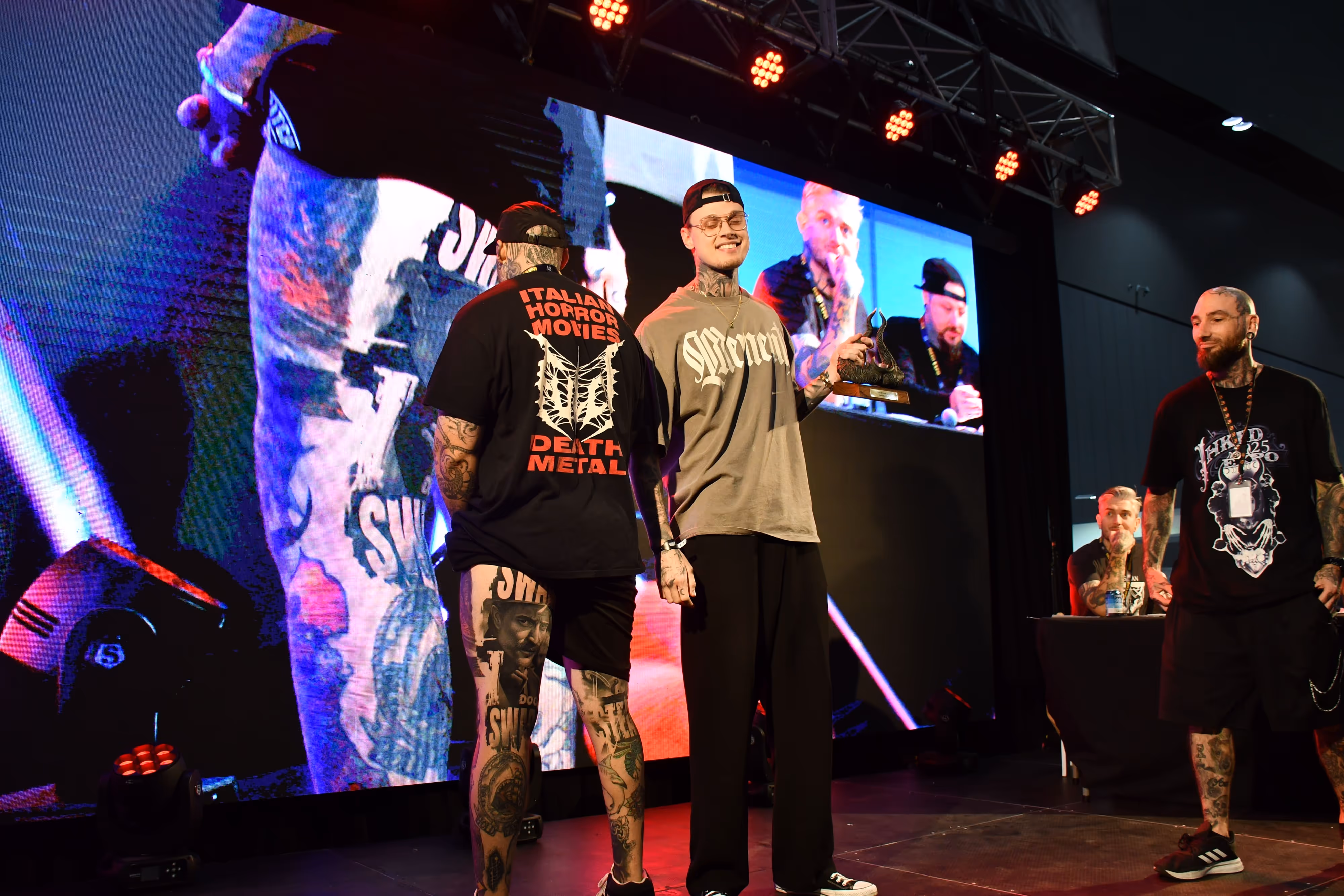Three tattooed men on stage at an event, one holding a trophy, with a large screen showing two men speaking in the background.