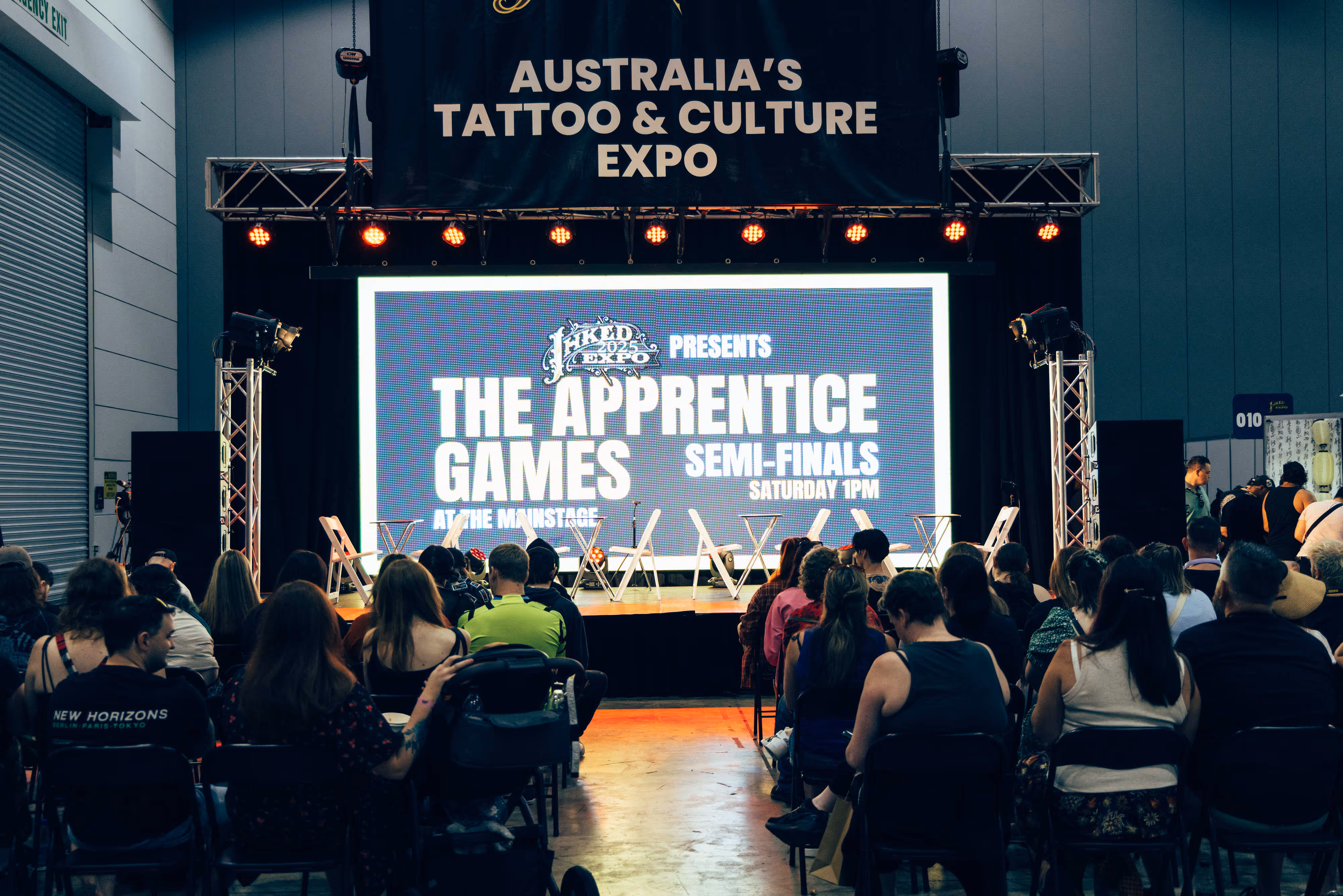 Audience seated facing a stage with a large screen announcing 'The Apprentice Games Semi-Finals Saturday 1PM' at Australia's Tattoo & Culture Expo.
