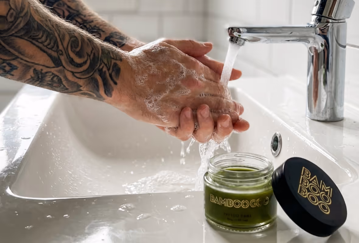 Tattooed hands washing with soap under running water from a chrome faucet beside an open jar of green BAMBOOCO skincare product on a white sink.