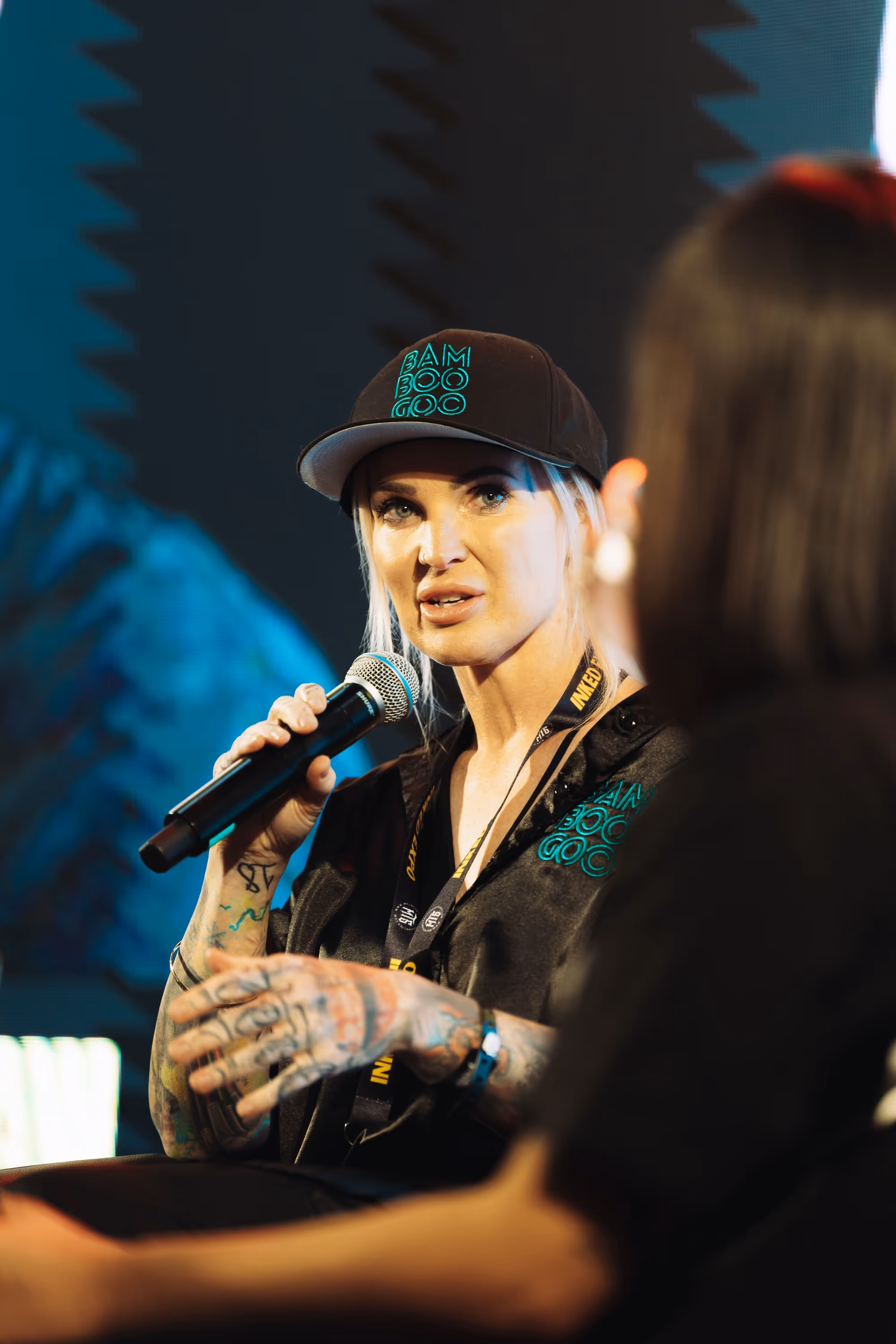Tattooed woman with blonde hair wearing a black cap and shirt speaking into a microphone during a discussion.