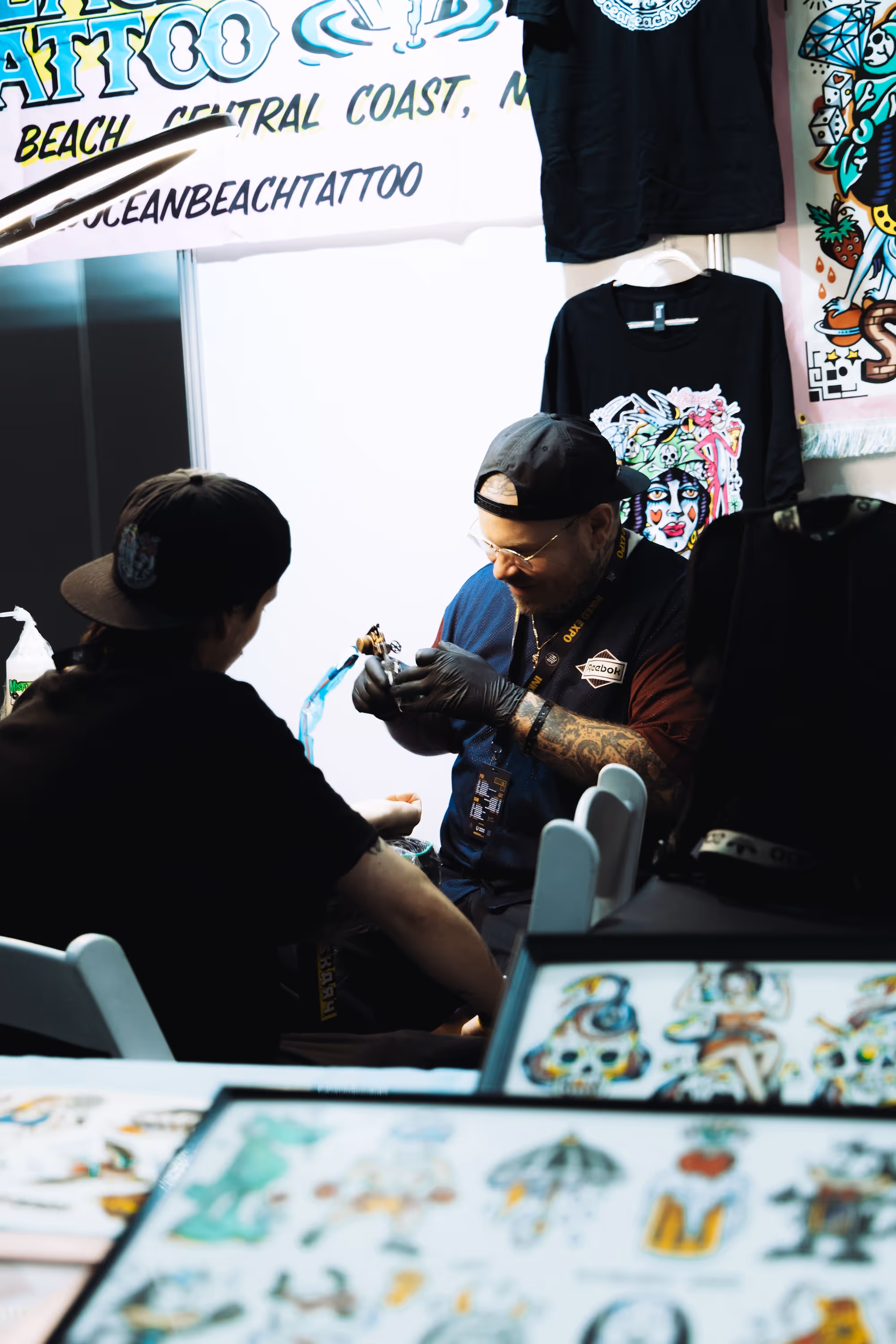 Tattoo artist wearing gloves and a cap tattooing a customer’s arm in a brightly lit booth with colorful artwork and T-shirts displayed.
