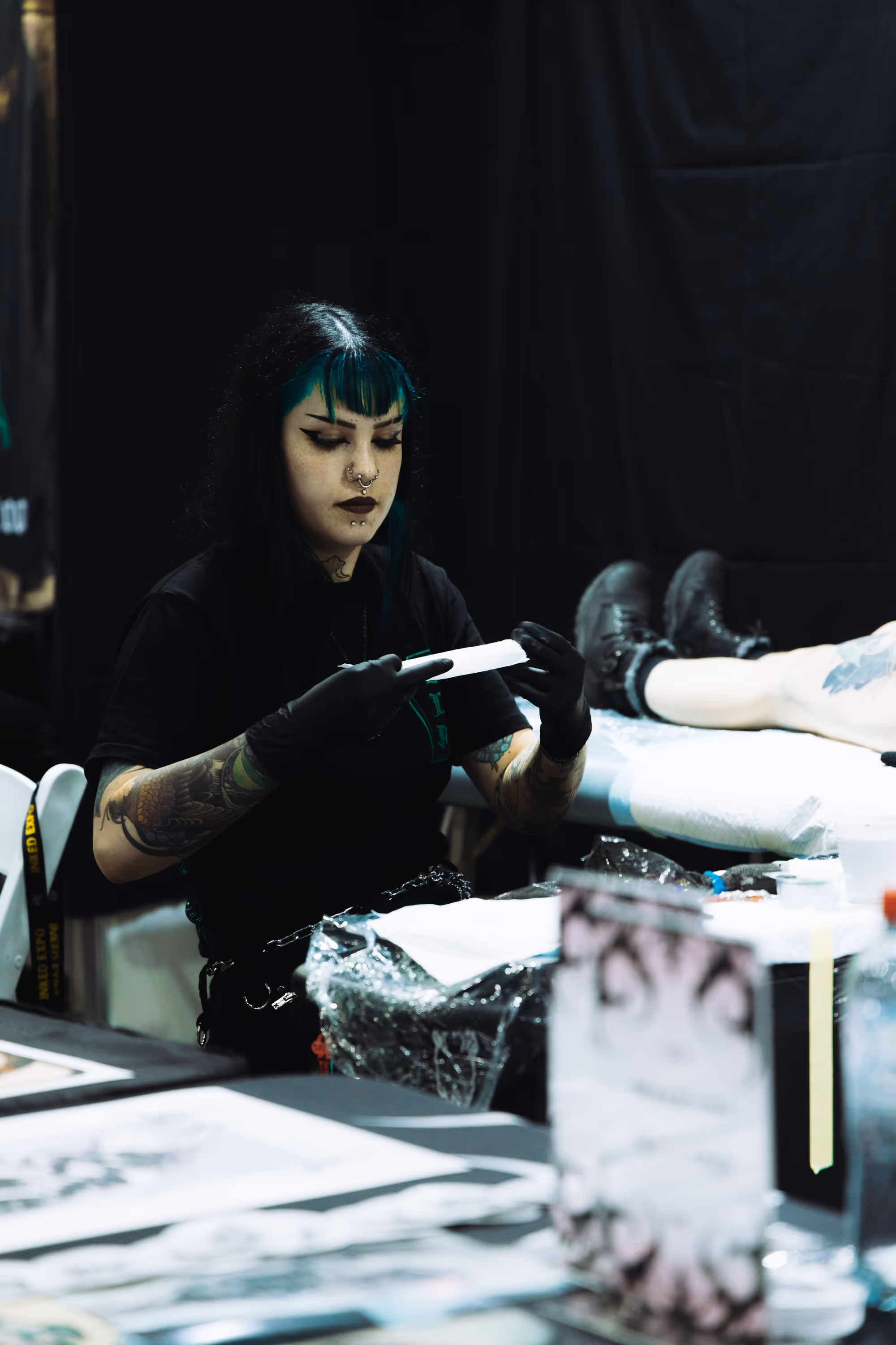 Tattoo artist with black gloves preparing a tattoo stencil while a client lies on a table in the background.