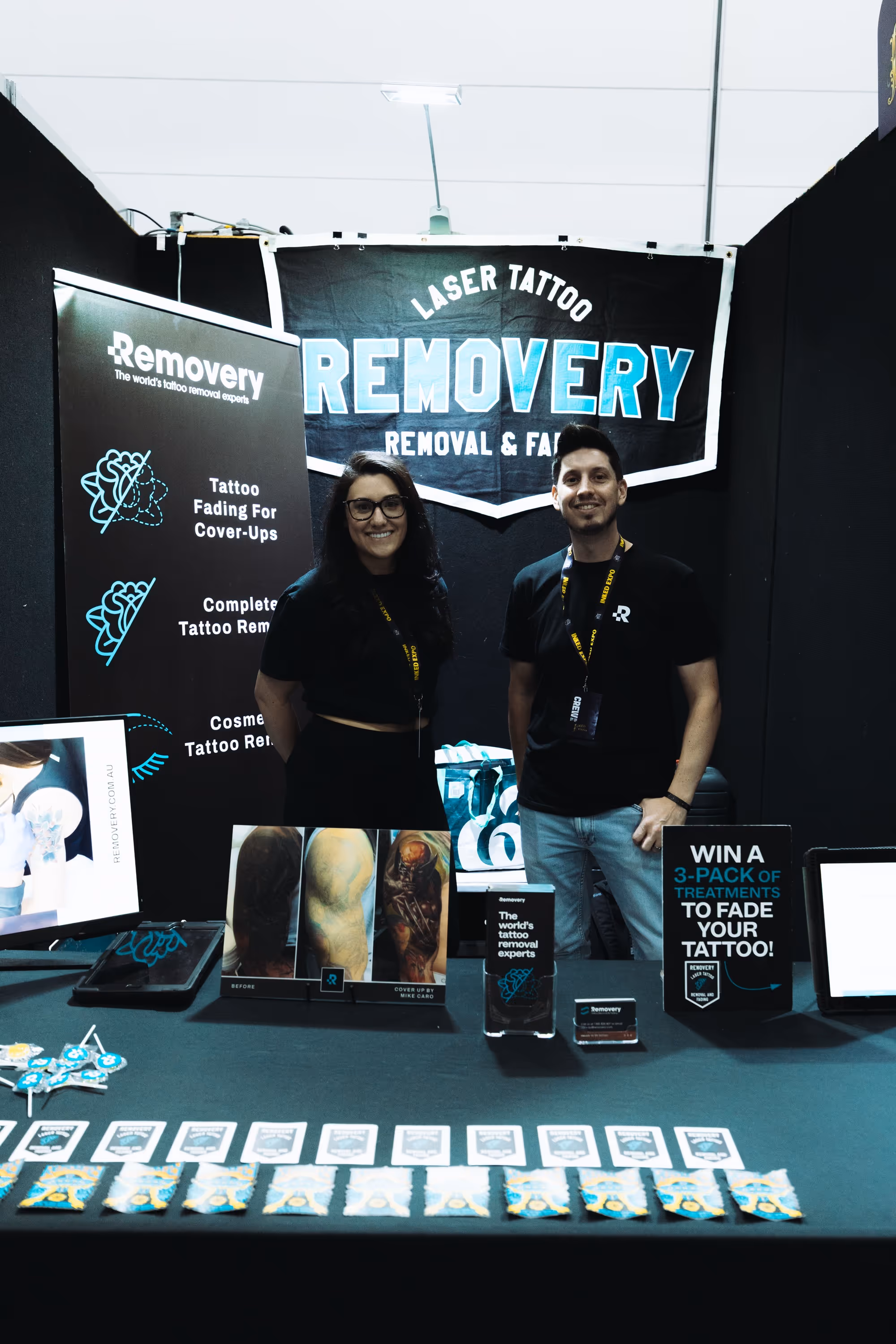 A man and woman standing behind a tattoo removal booth table with promotional items and displays at an event.
