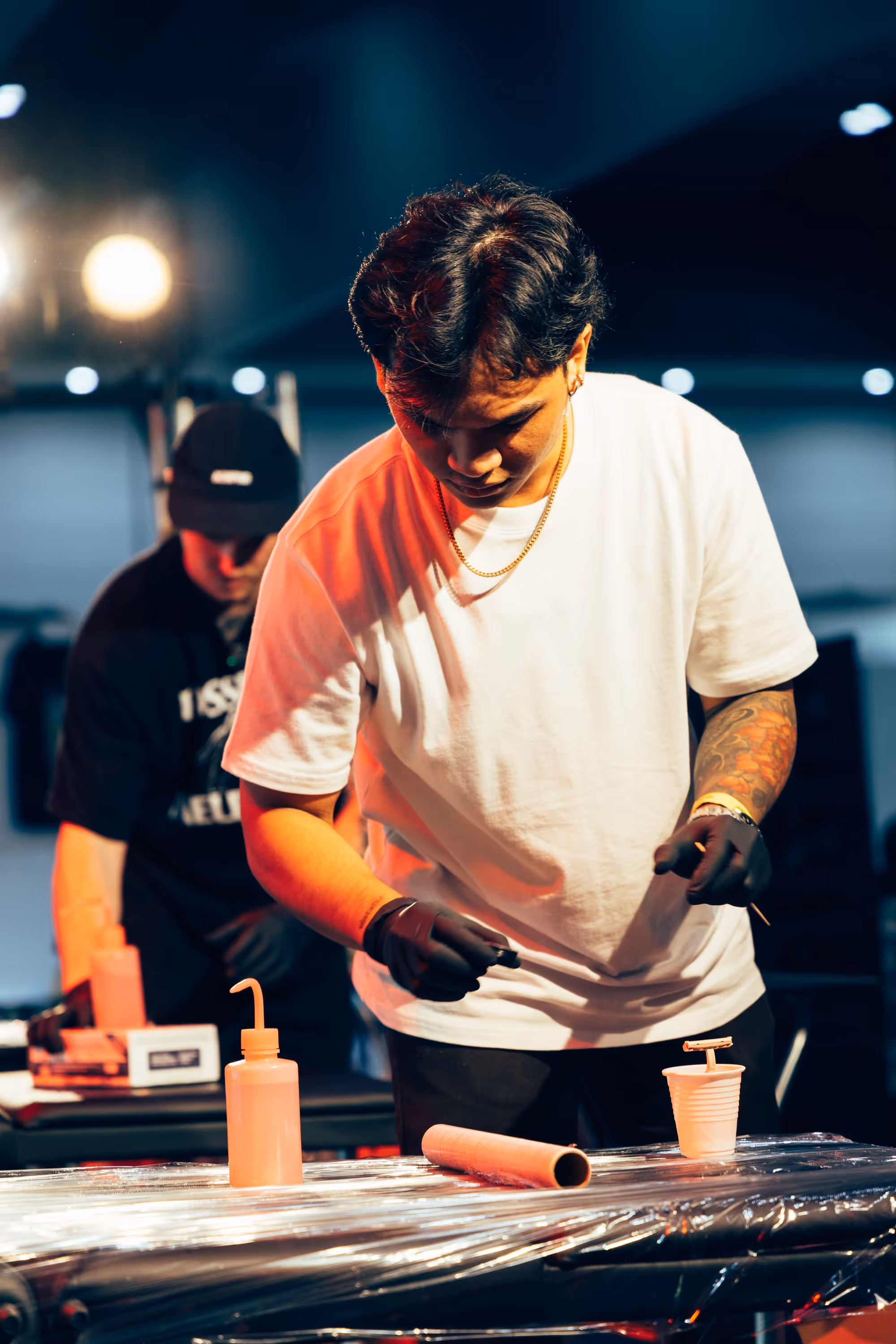 Man in white t-shirt with tattooed arm and black gloves working at a table with a plastic cup and squeeze bottle under warm lighting.
