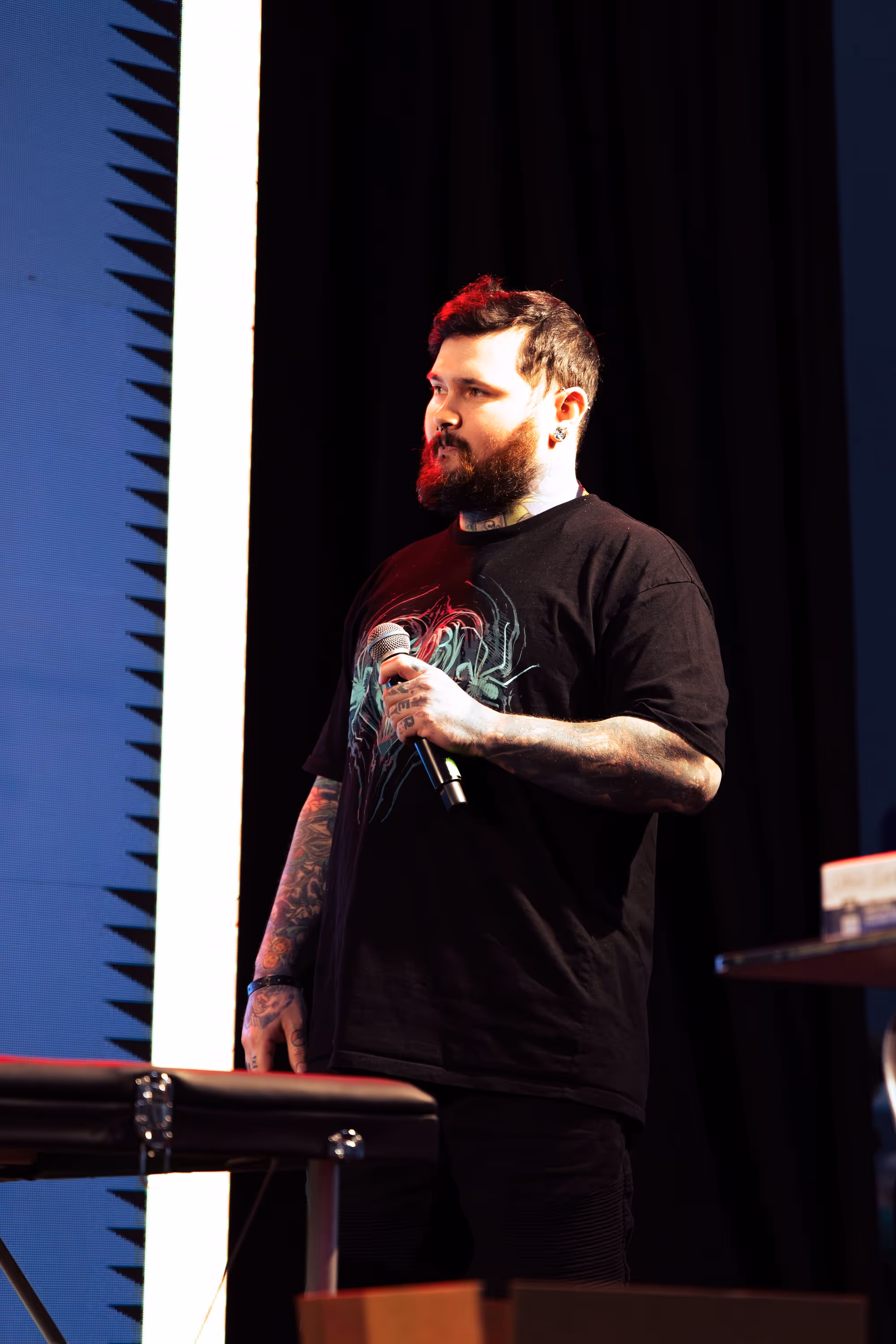 Bearded man with tattoos holding a microphone on stage with a dark background.
