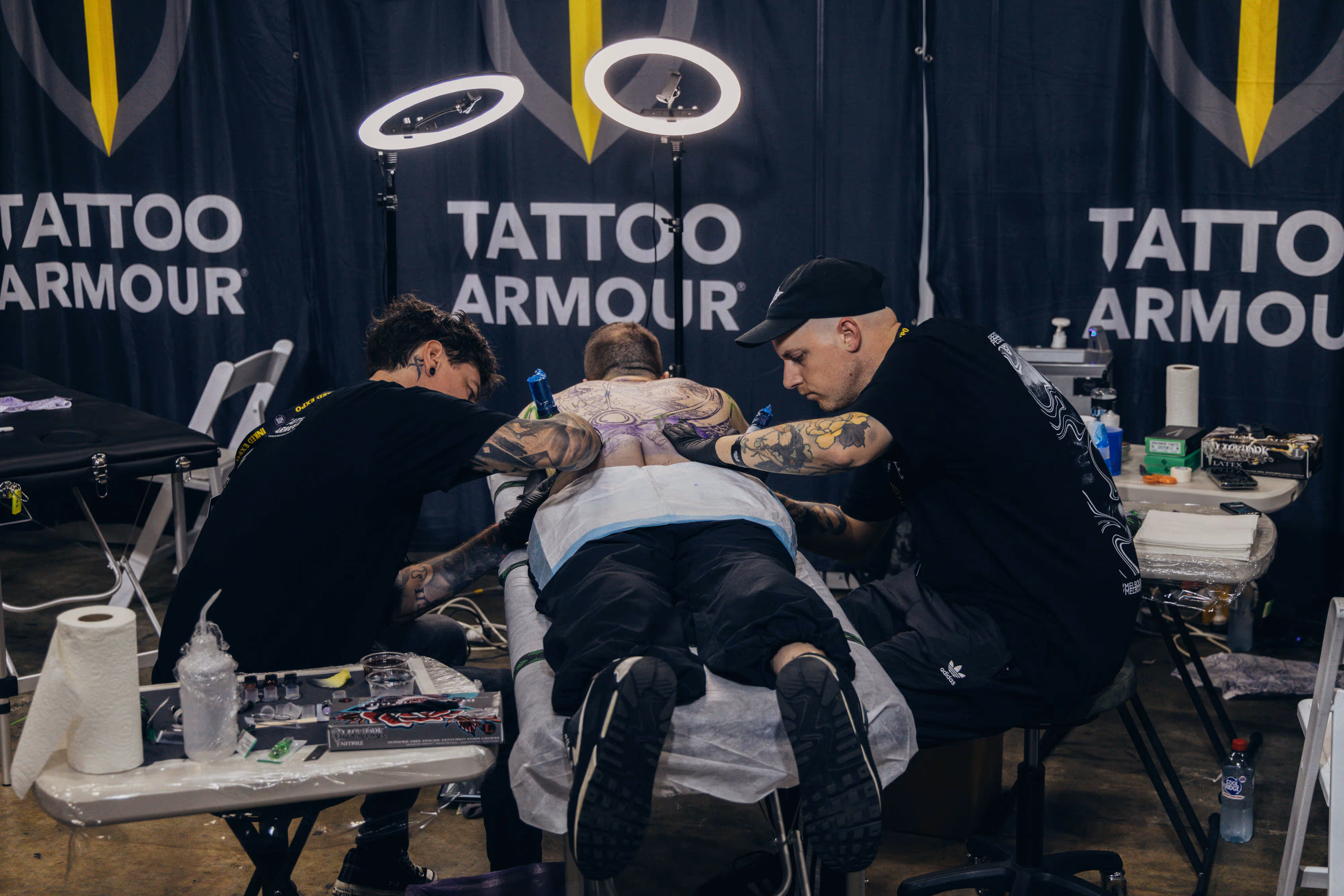 Two tattoo artists working on a large back tattoo of a person lying face down on a tattoo table under ring lights.