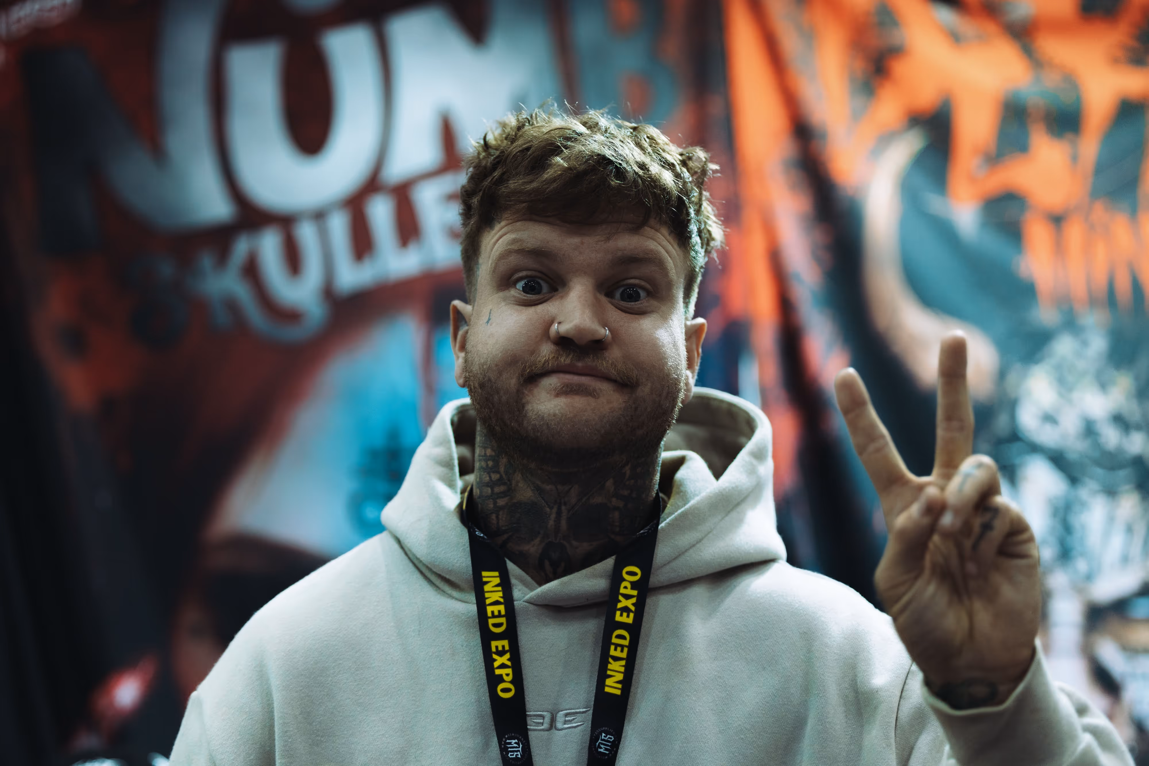 Man wearing a light hoodie and Inked Expo lanyard making a peace sign with his fingers in front of a colorful graffiti wall.
