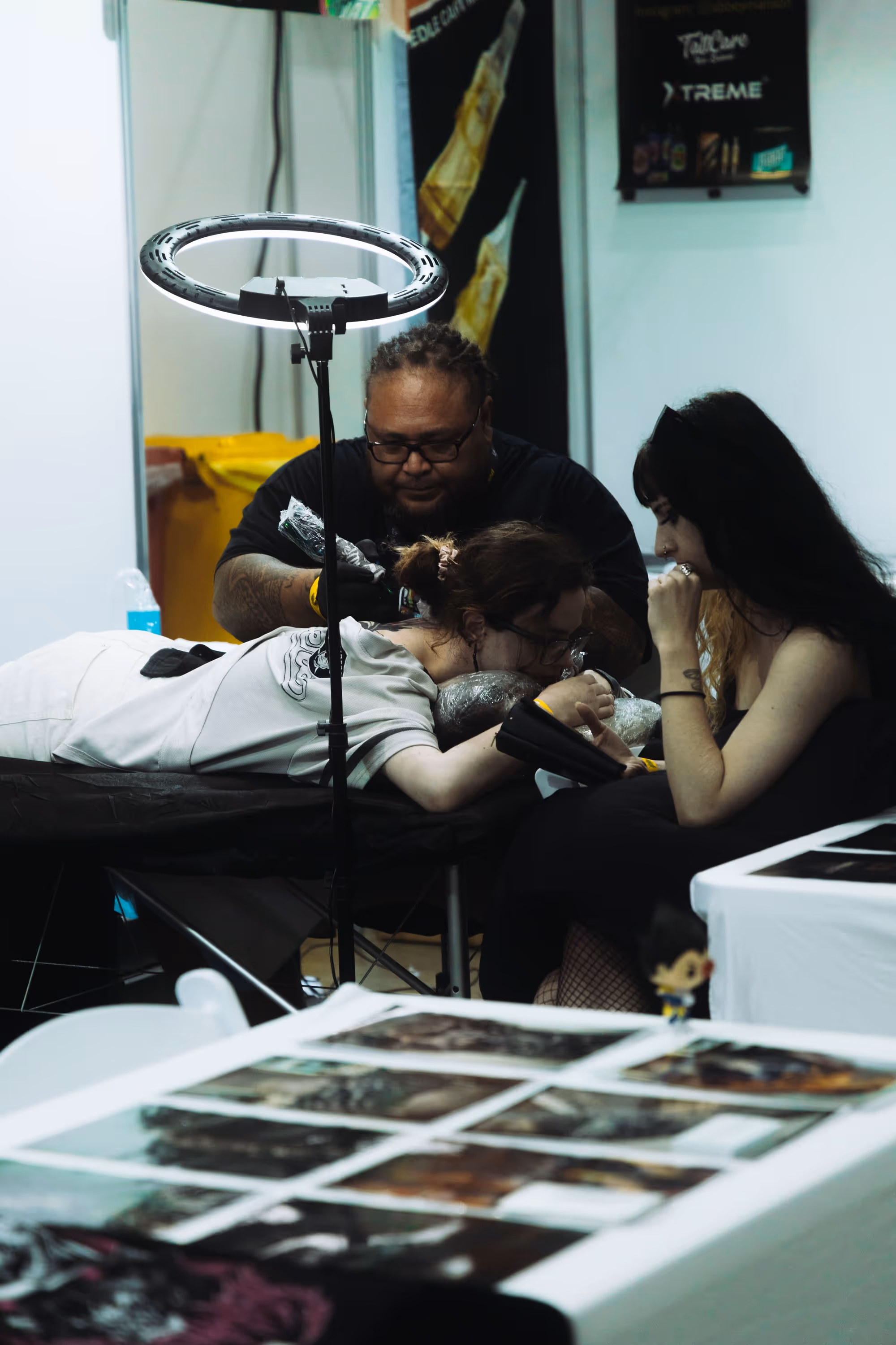 Tattoo artist with dreadlocks and glasses working on a client lying face down on a table while another woman watches.