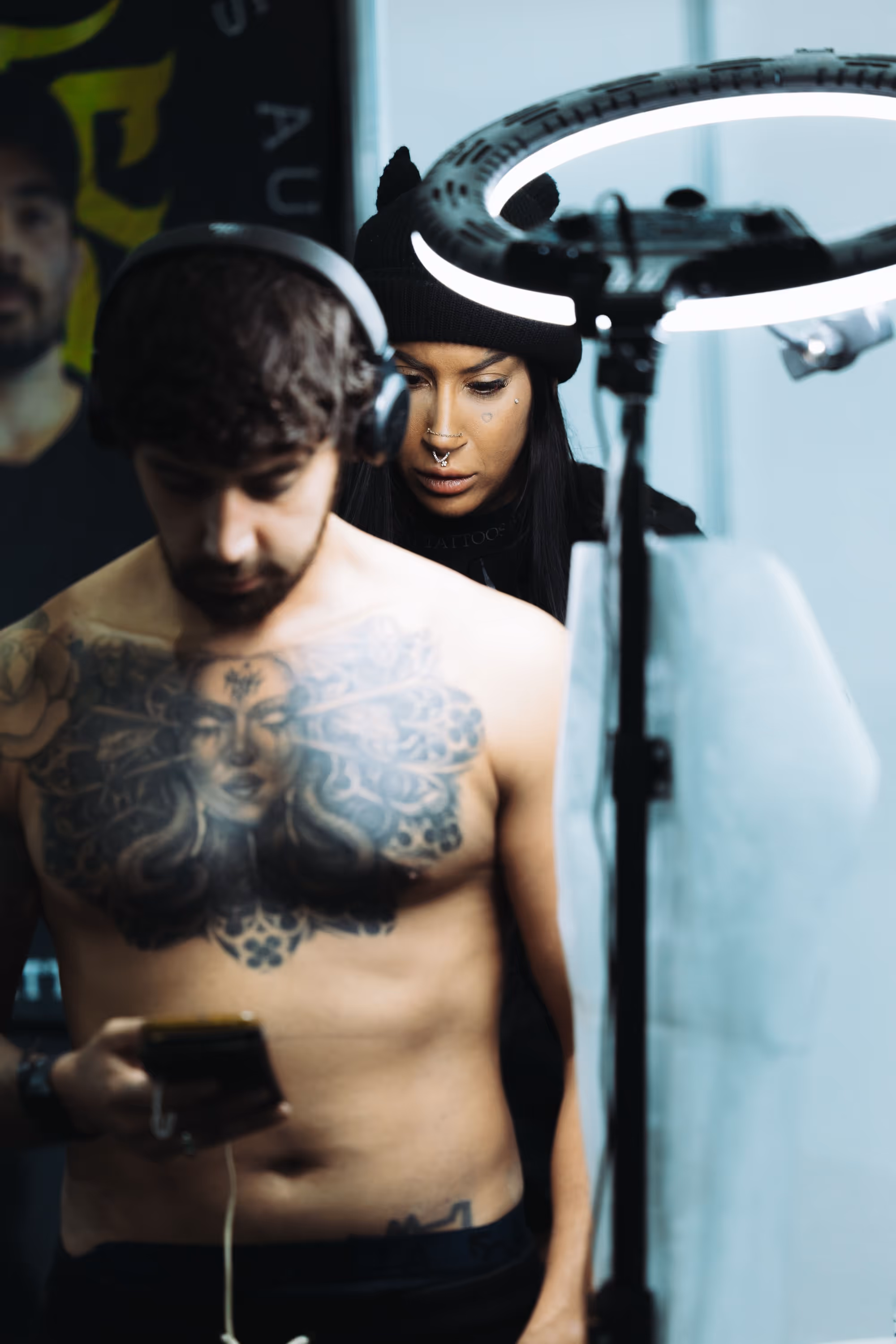 A shirtless man with a large chest tattoo looks at his phone while a woman wearing a black beanie and facial piercings works behind him under a ring light.