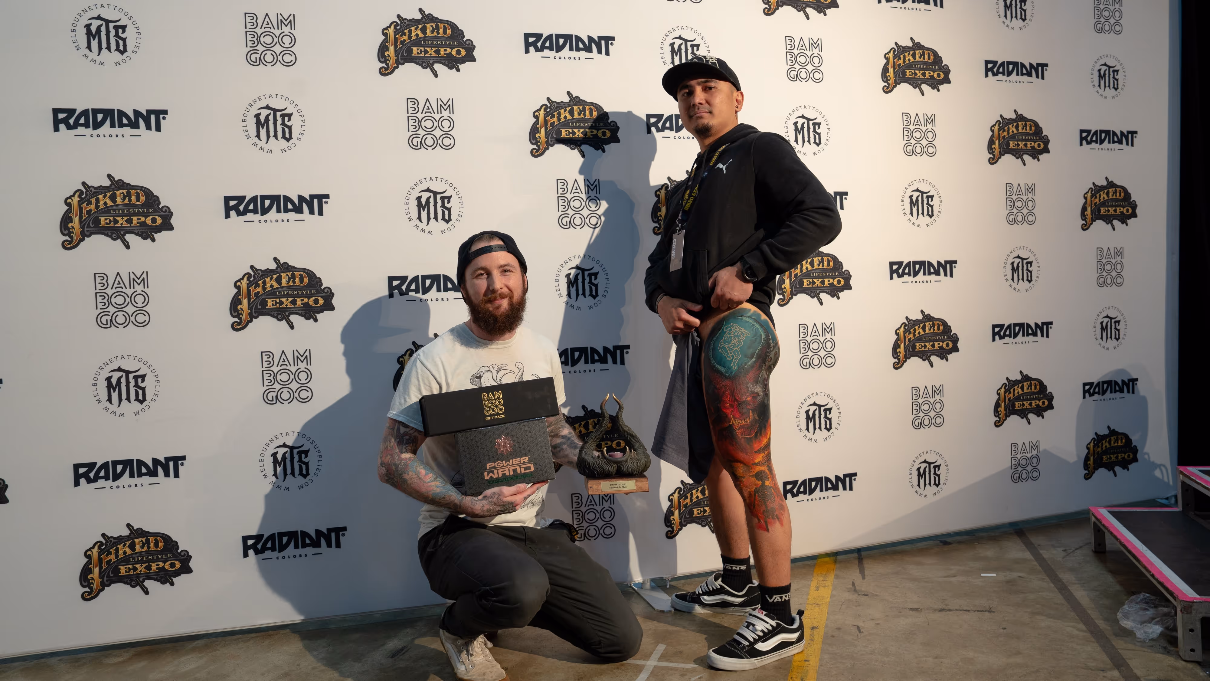 Two men posing in front of a banner with tattoo-related logos, one kneeling holding two award boxes and a trophy, the other standing showing a colorful thigh tattoo.