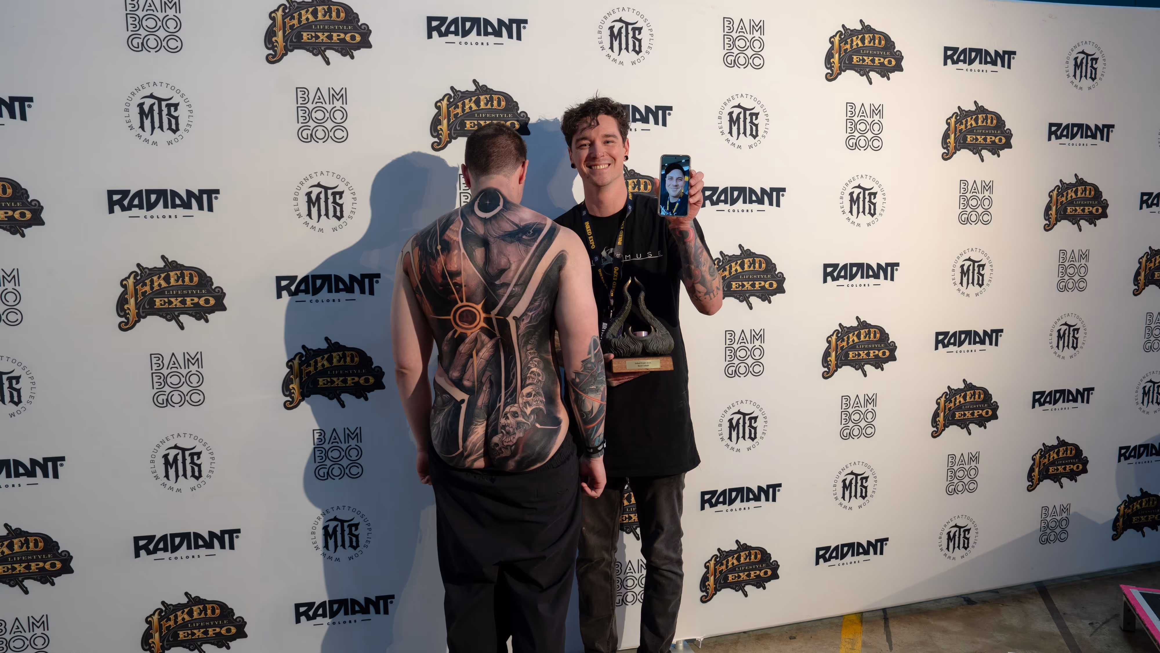 Two men at a tattoo expo, one displaying a detailed full-back tattoo and the other holding a trophy and smartphone showing a selfie.