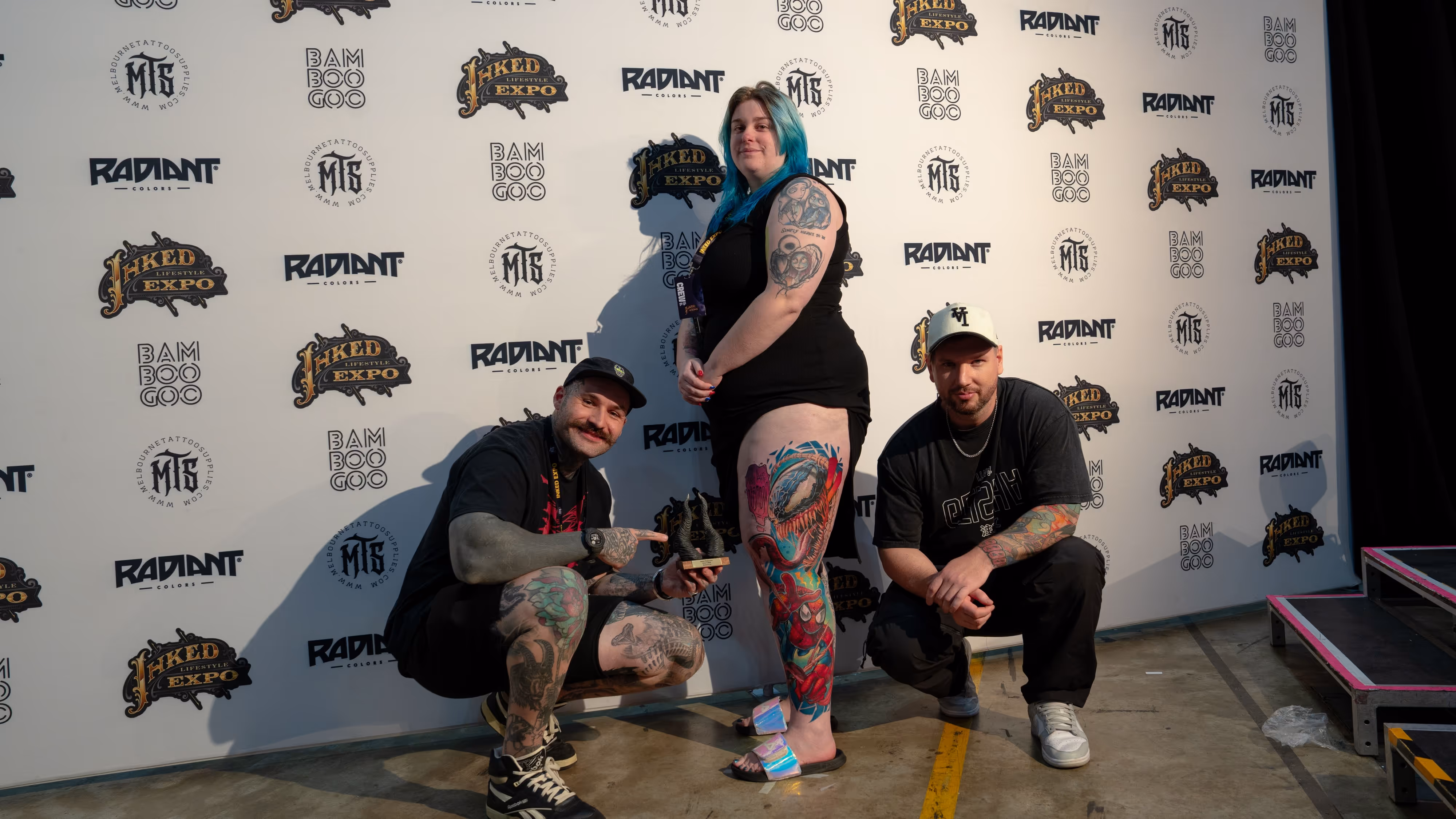 Three tattooed people posing at Inked Lifestyle Expo, with a woman showing a colorful leg tattoo featuring Spider-Man and a shark.