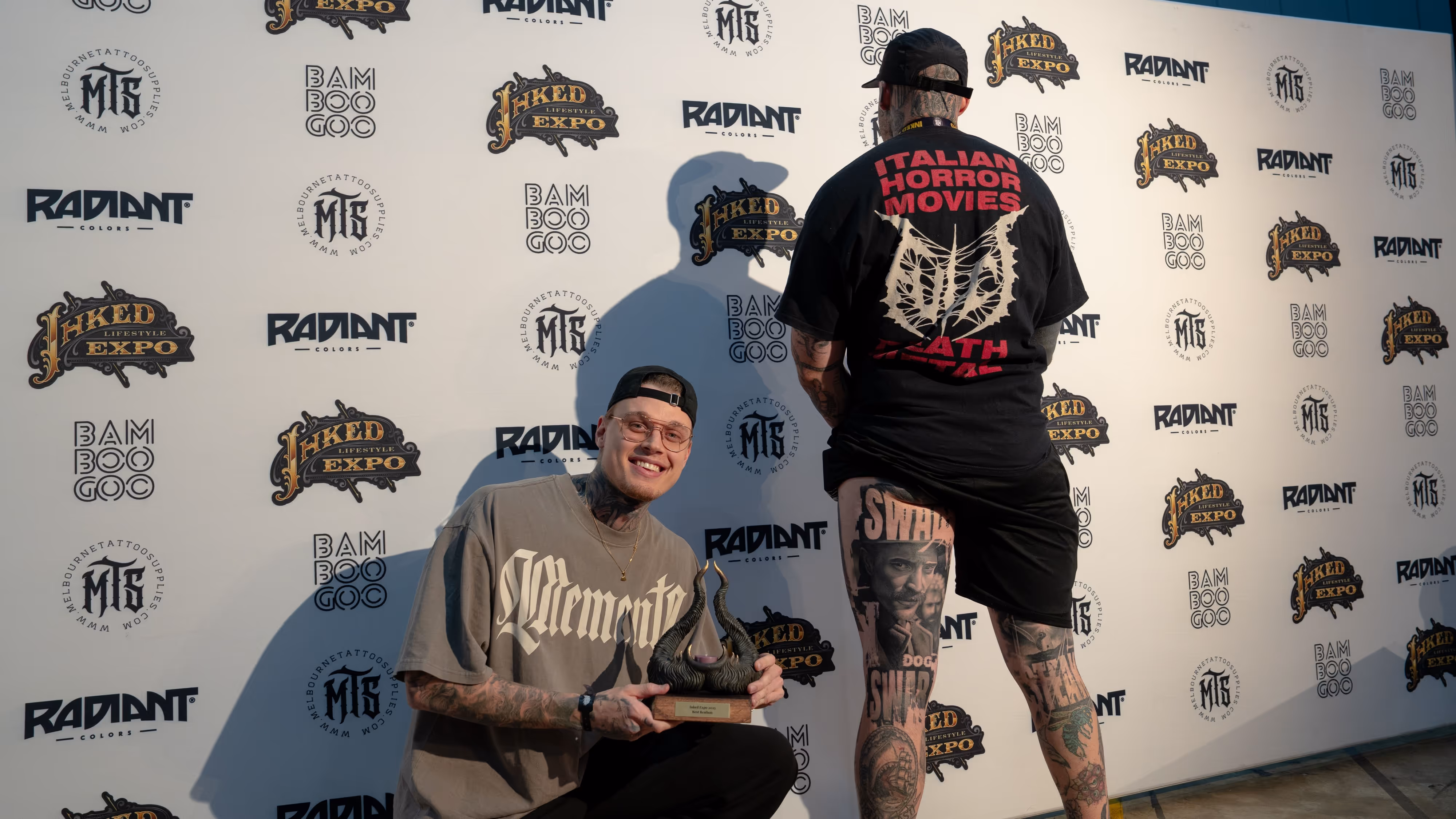 Two tattooed men posing in front of a backdrop with event logos; one crouches holding a horned trophy while the other shows tattoos on his leg and back.