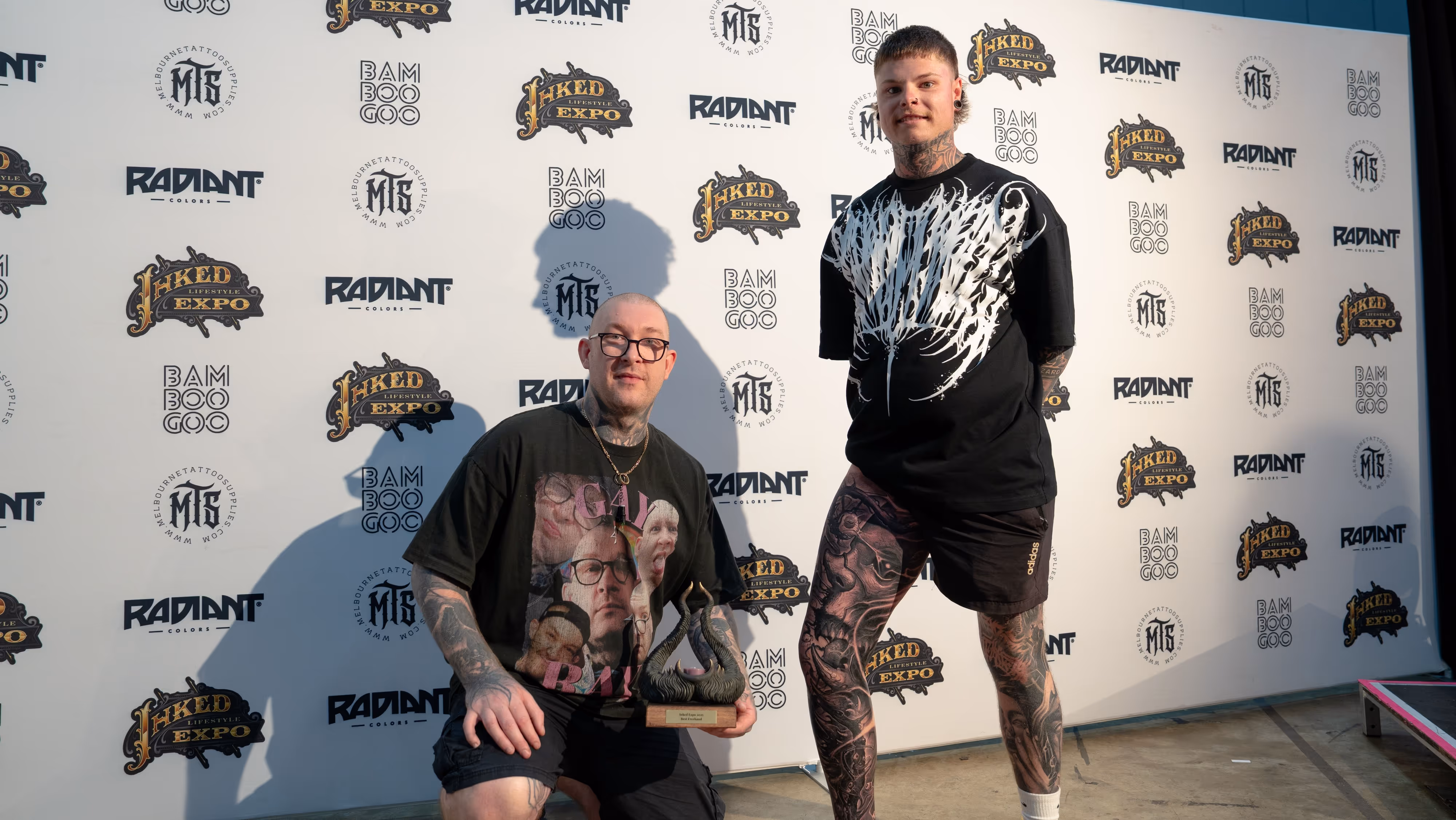 Two tattooed men posing in front of a backdrop with Inked Expo and Radiant logos; one man is holding a trophy.