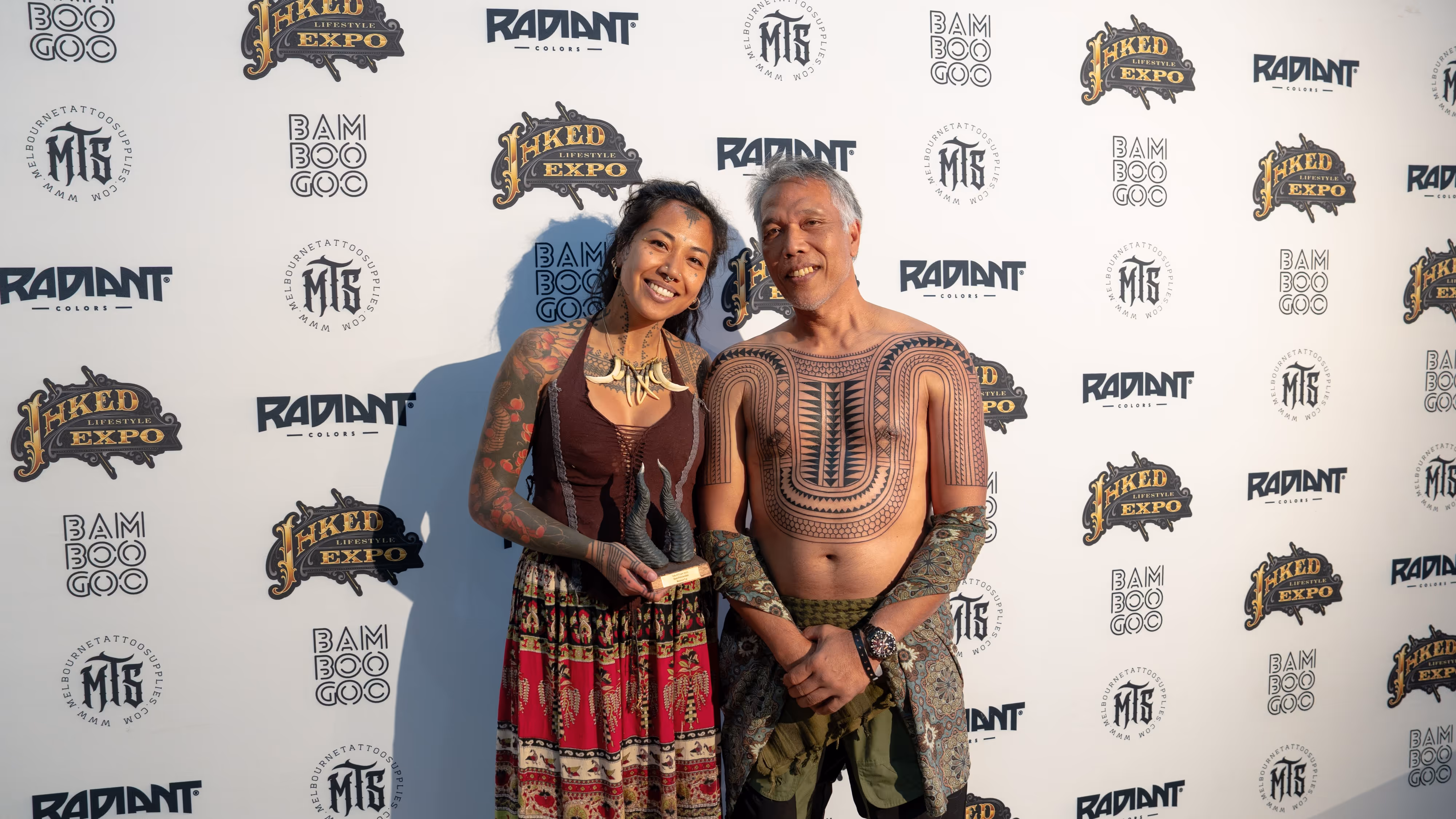 Smiling tattooed man and woman standing in front of a lifestyle expo backdrop, with the woman holding a horned trophy.