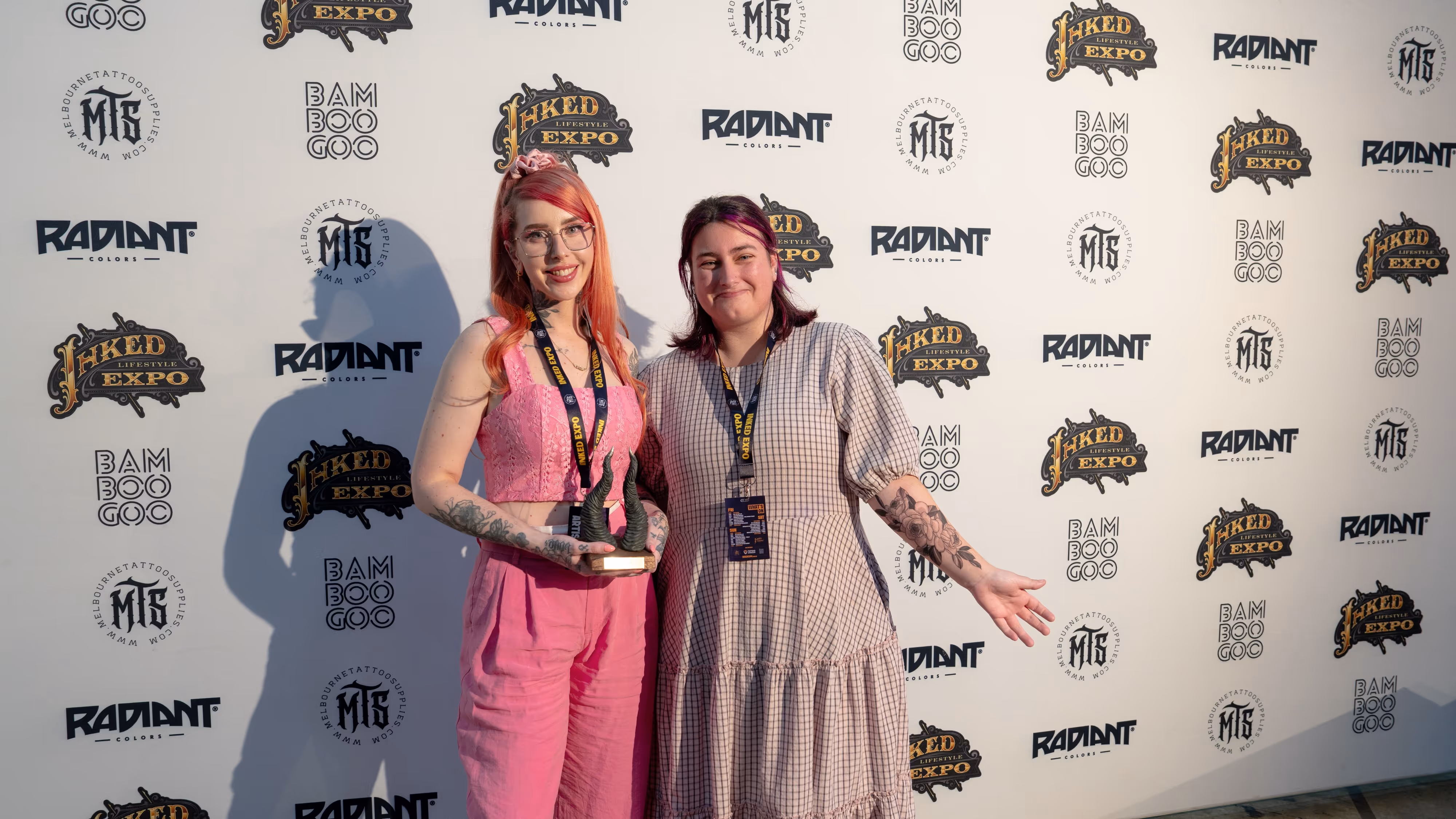 Two smiling women with tattoos posing in front of a branded backdrop at Inked Expo, one holding a trophy with horn-shaped design.