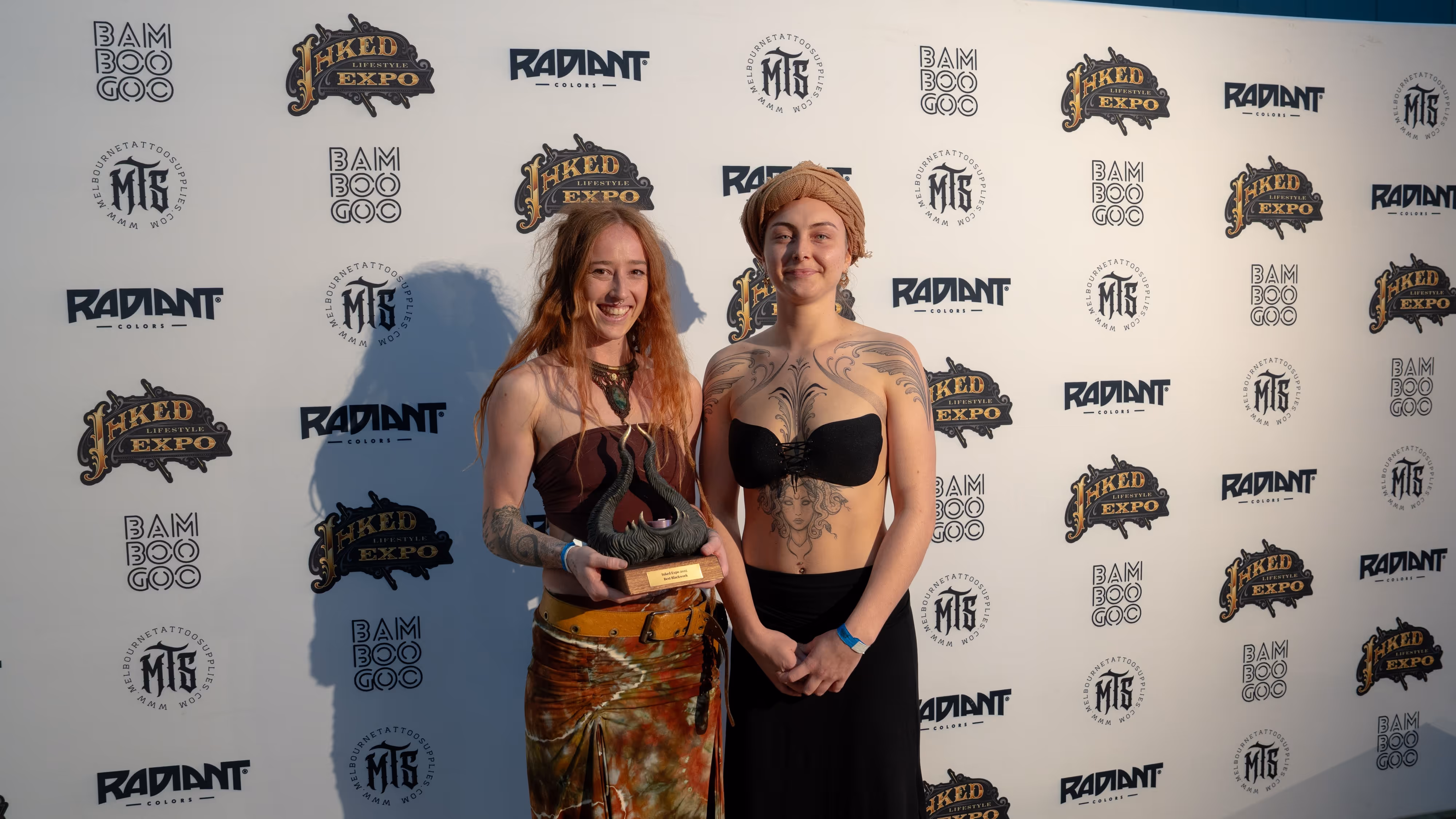 Two women standing in front of a backdrop with sponsor logos, one holding a trophy and both wearing strapless tops and skirts, showcasing tattoos.