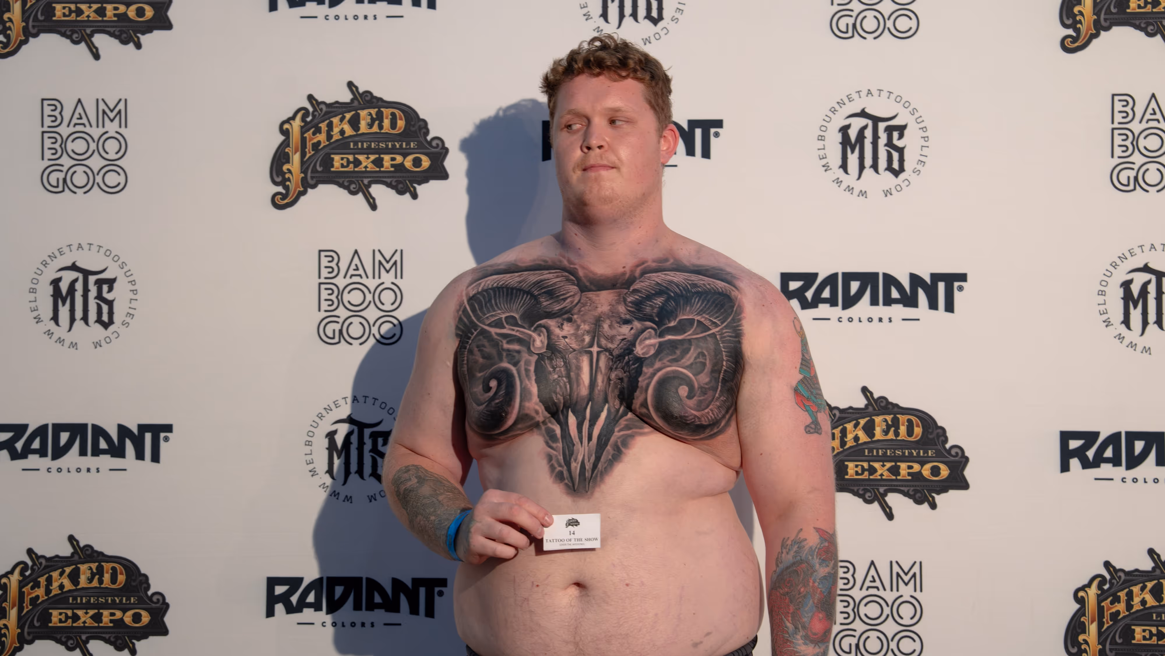Man with a large black ram skull tattoo covering his chest holding a 'Tattoo of the Show' card at a tattoo expo backdrop.