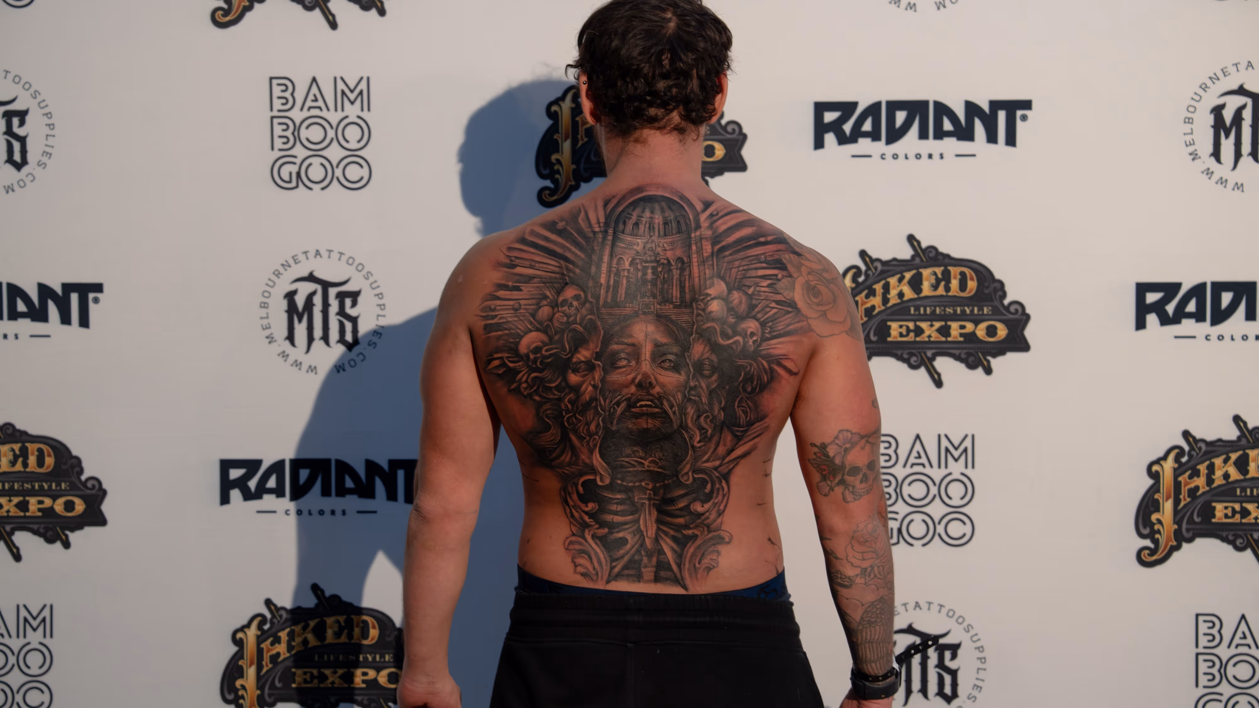 Man with a detailed black and gray back tattoo featuring skulls, a central face, and architectural elements, standing against a white background with multiple tattoo expo logos.