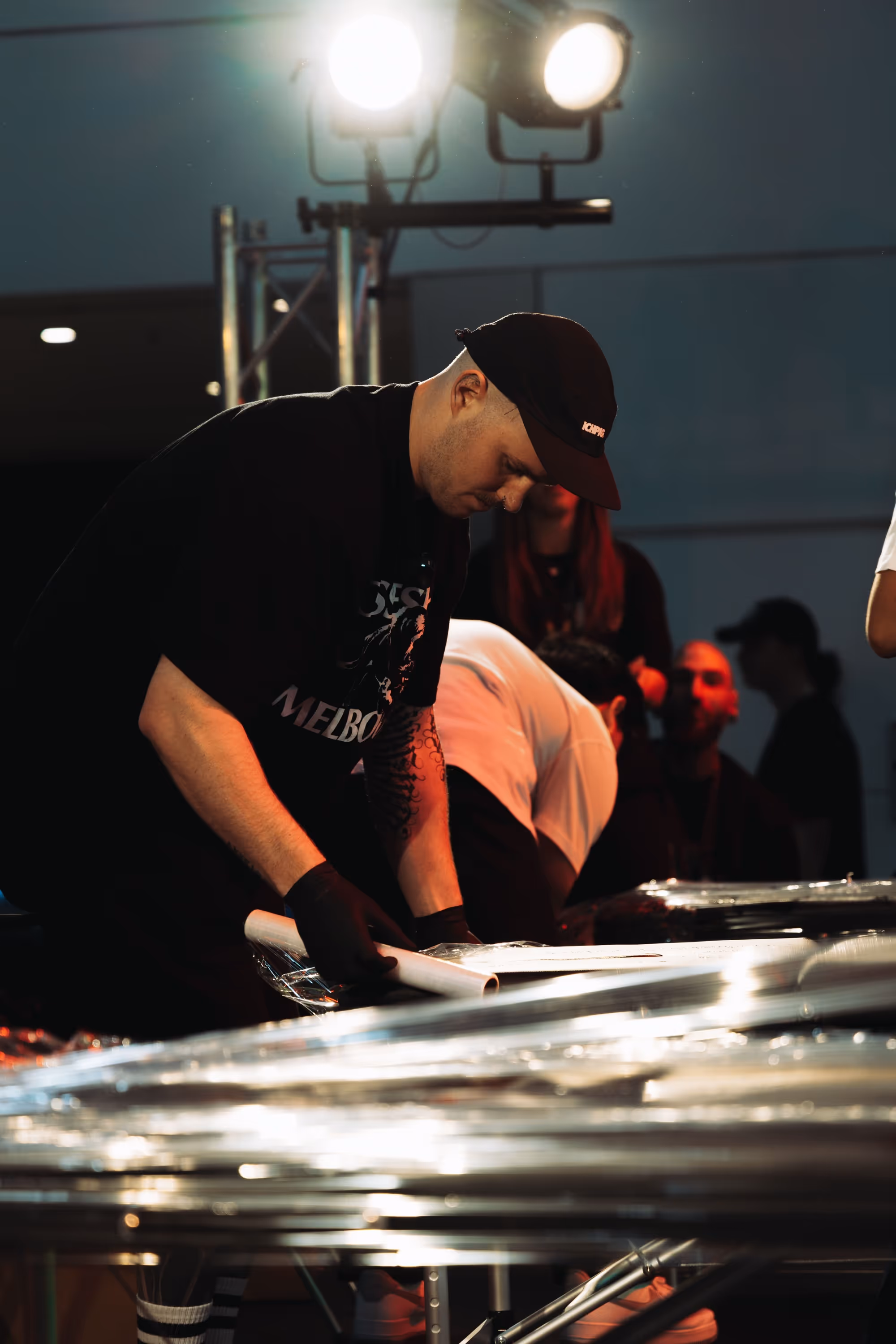 Man wearing a black cap and gloves working with rolled-up papers under bright stage lights.
