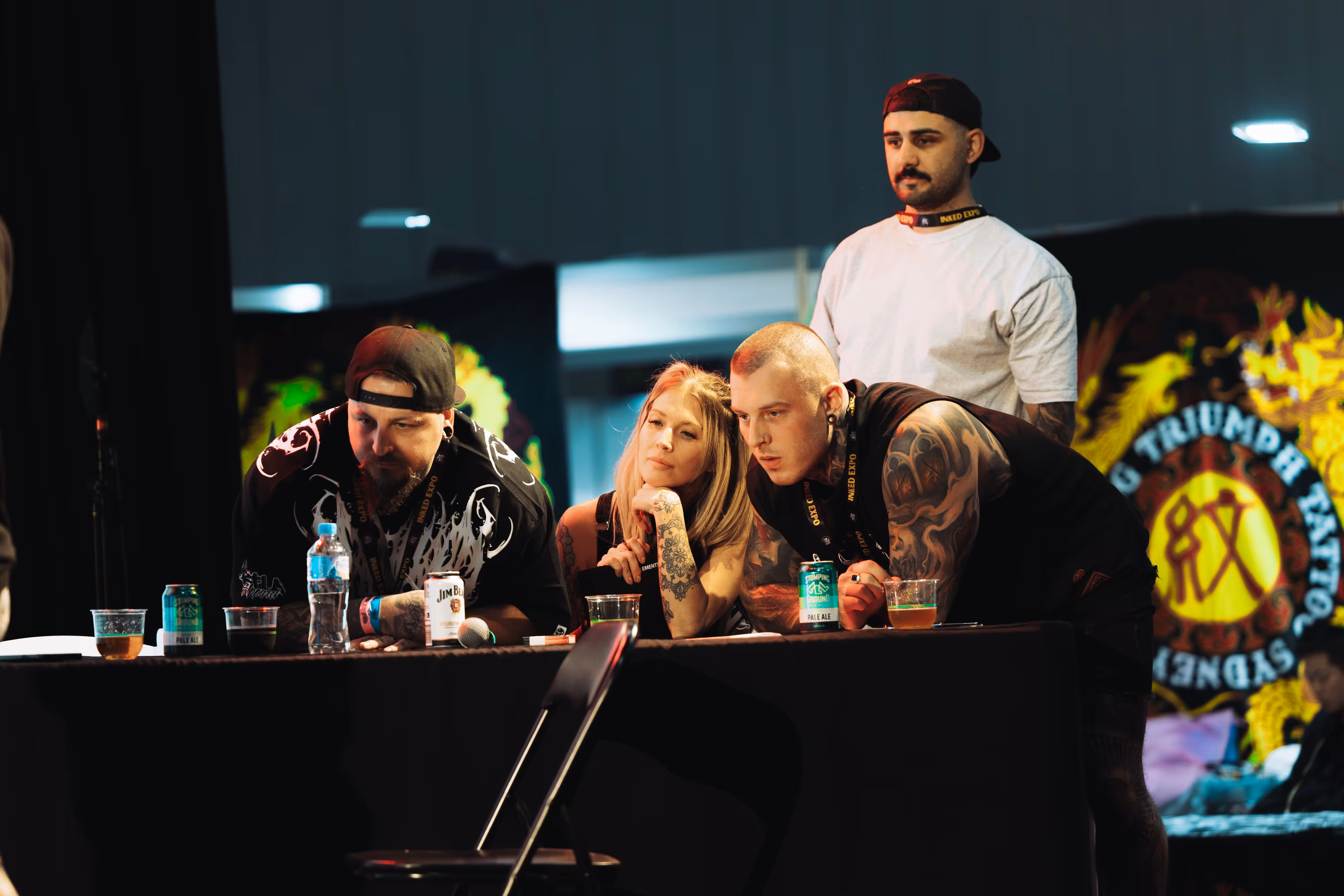 Four tattooed people, three leaning on a table with drinks, attentively watching something at a tattoo expo.