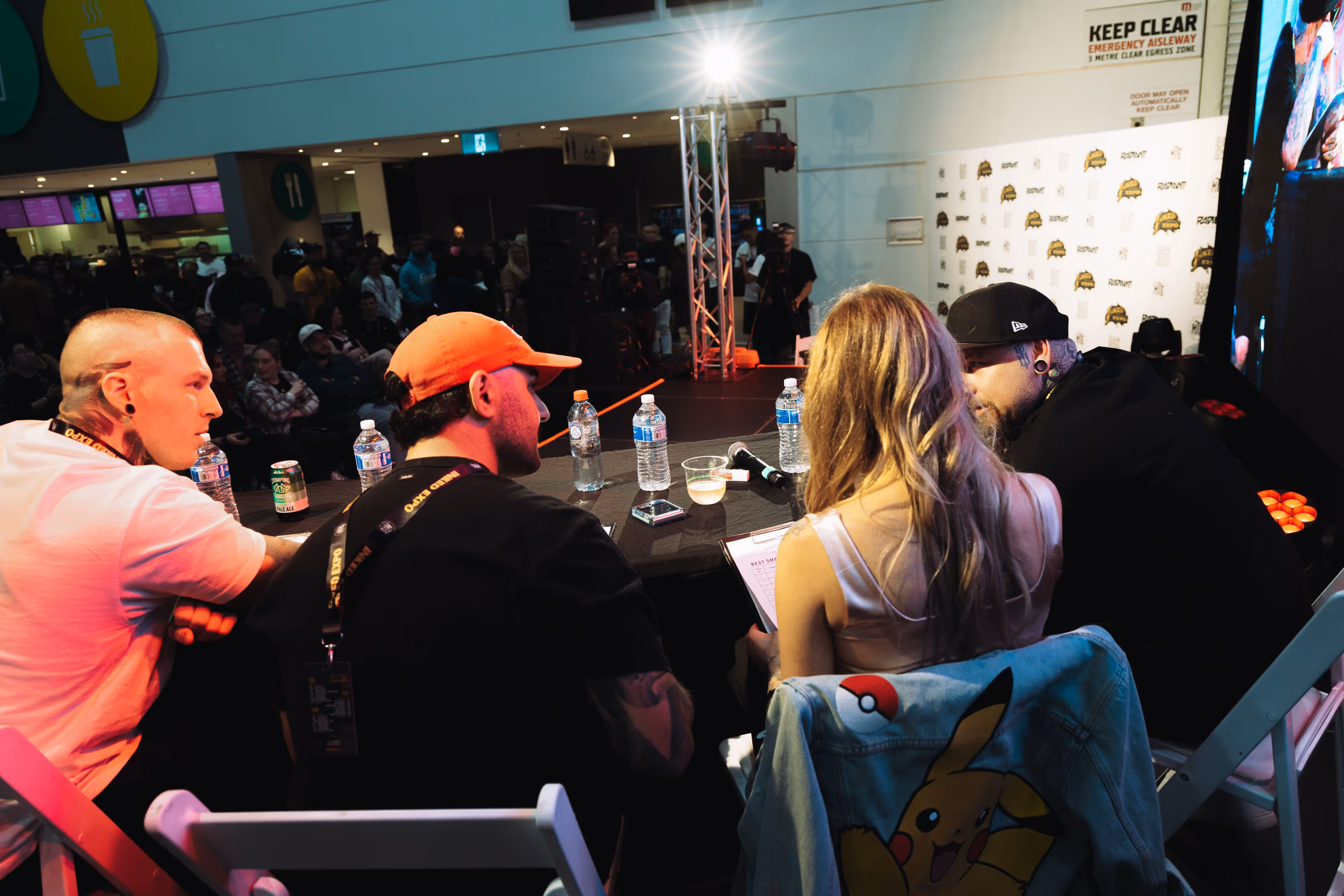 Four judges seated at a table with drinks and a microphone, facing an audience at an event.
