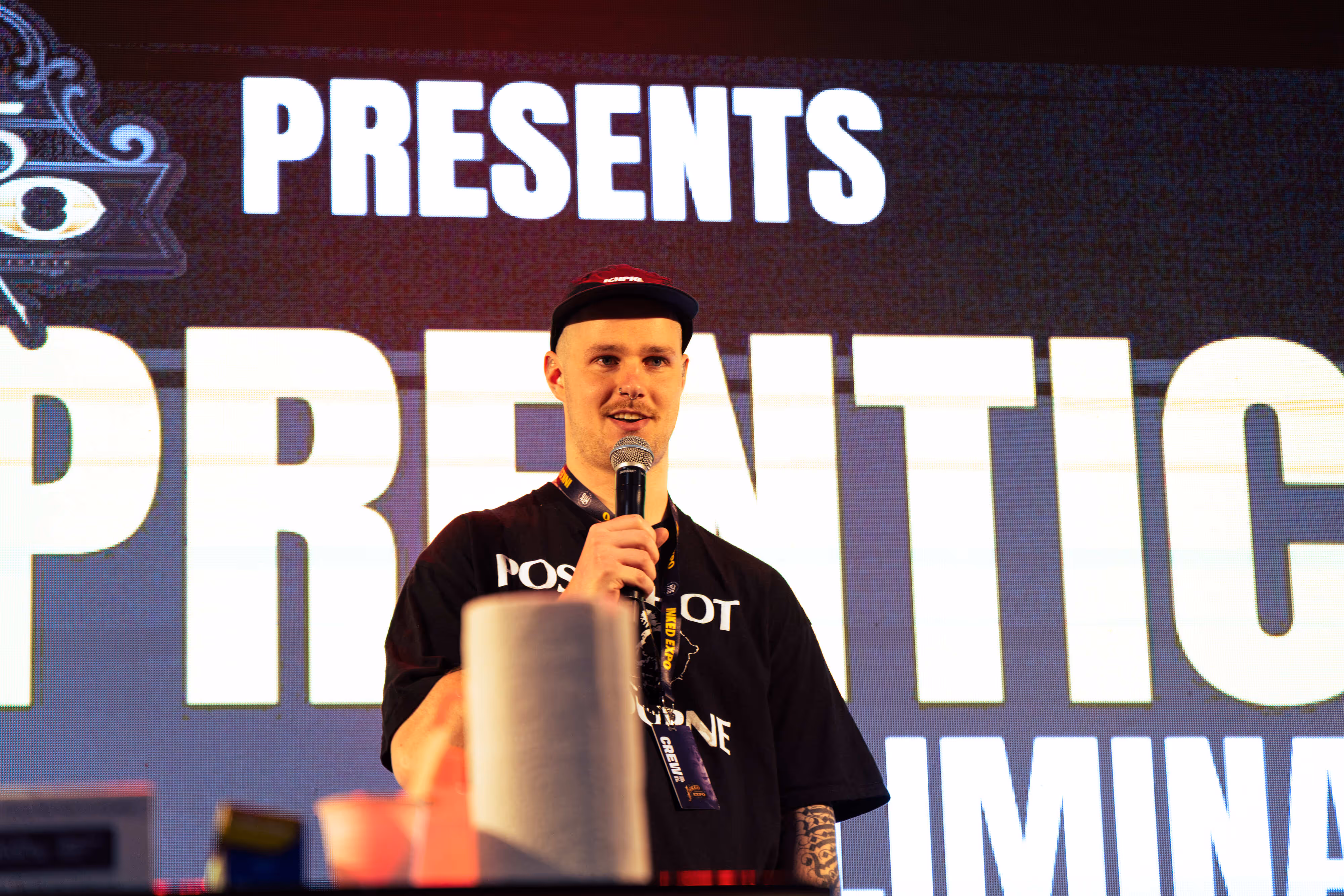 Man with a mustache and cap speaking into a microphone in front of a large illuminated screen displaying the word PRESENTS.