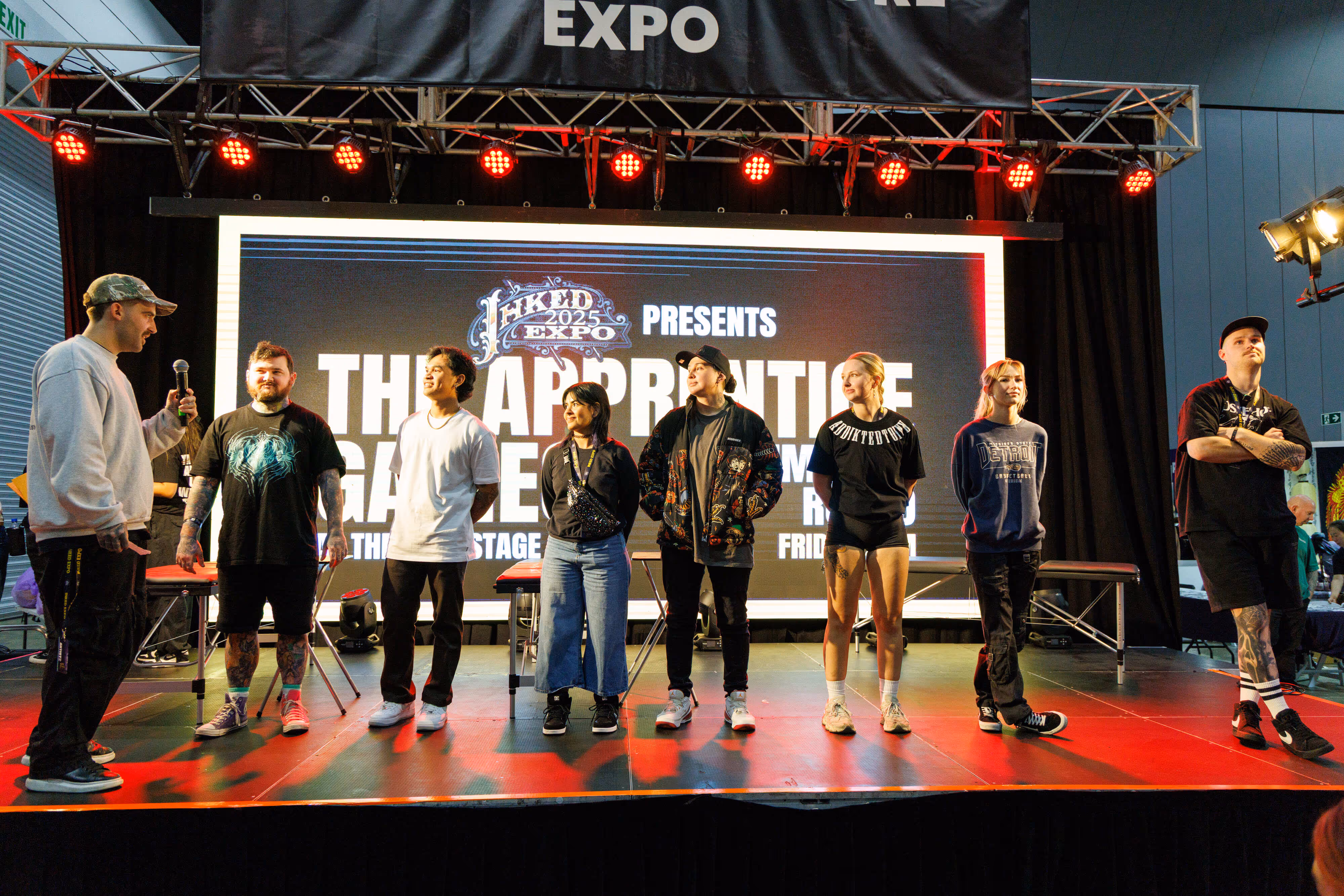 Eight people standing on a stage at an expo event, with a large screen behind them displaying 'TATTOO EXPO 2025' and event details.